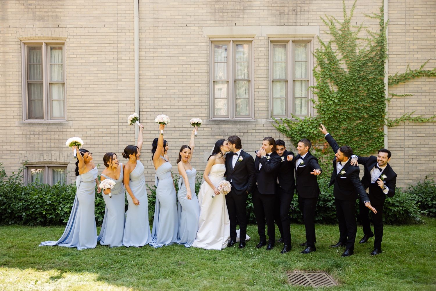Wedding party in light blue dresses and black tuxedos poses together on grass near brick building.