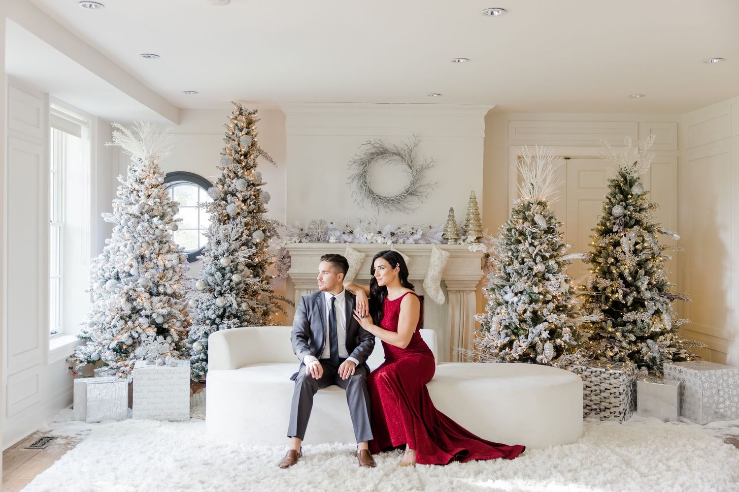 Festive holiday portrait with couple on white bench surrounded by snow-covered Christmas trees and mantel decor.