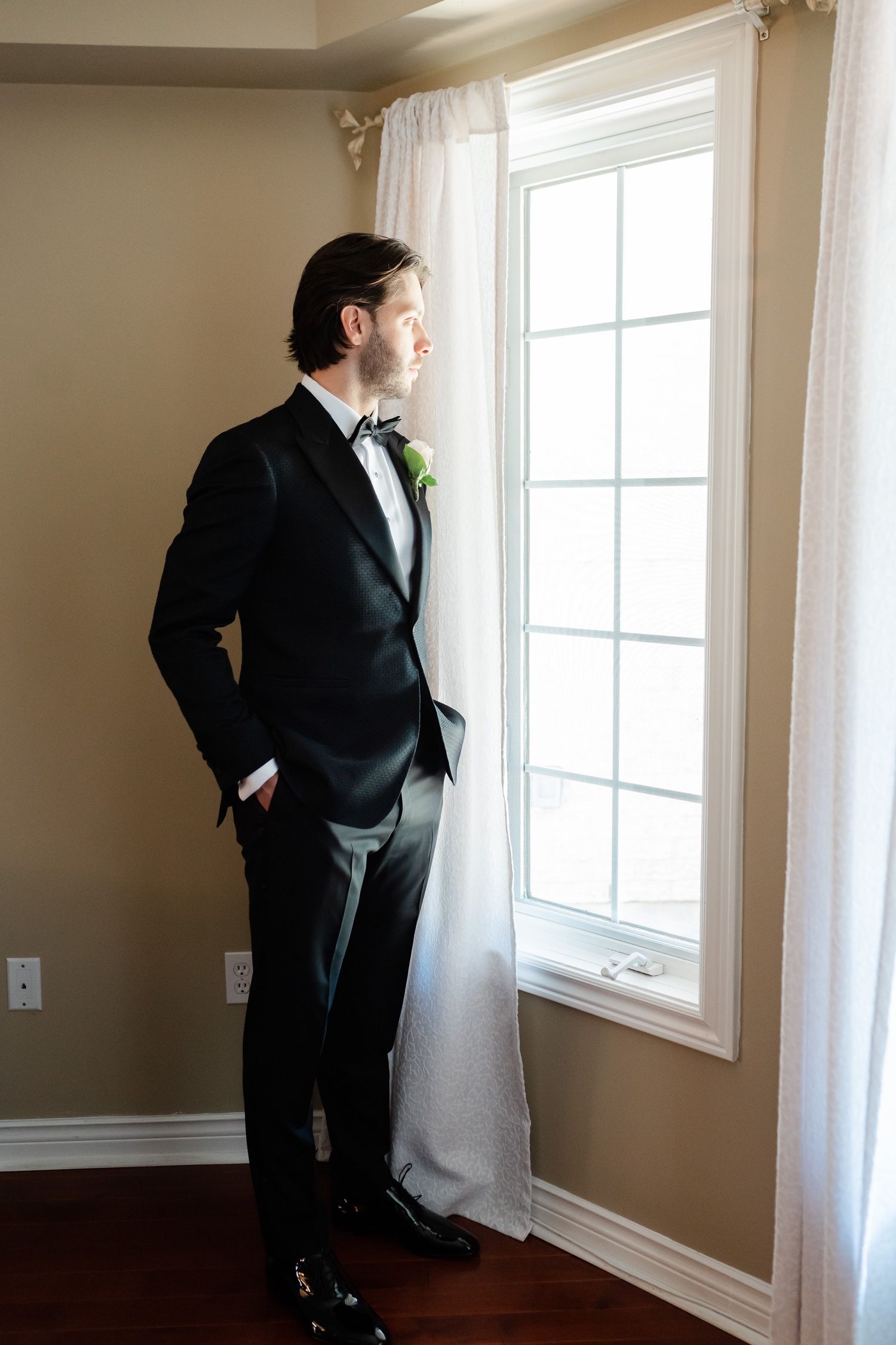 A formal portrait of a person in tuxedo standing by window with sheer white curtains.