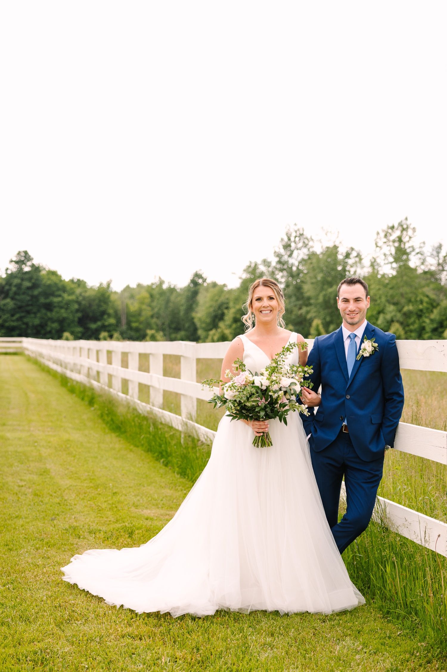 Wedding couple poses beside a white fence with a beautiful wildflower bouquet.
