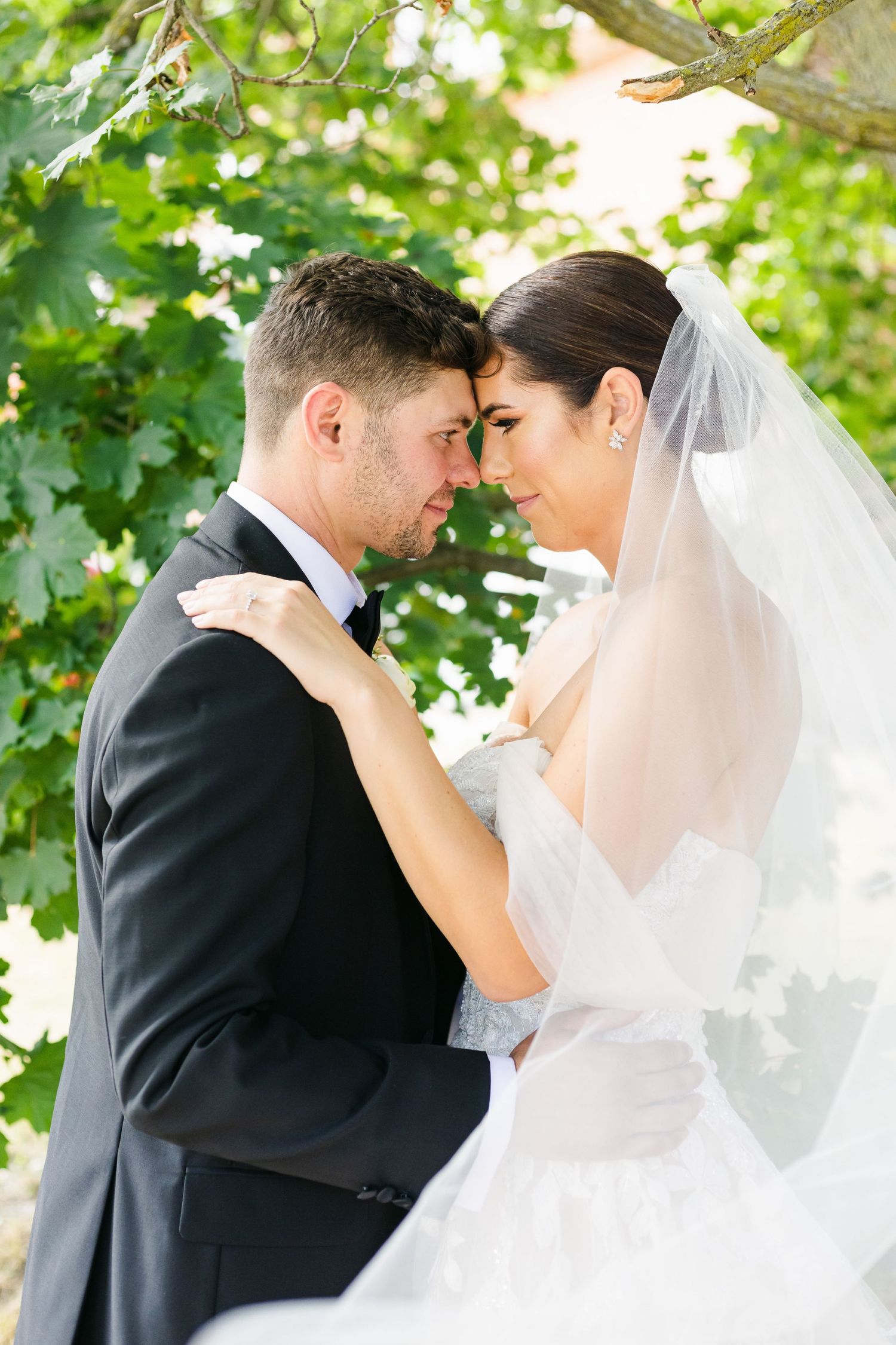 Intimate wedding moment captured under green leaves with flowing white veil.