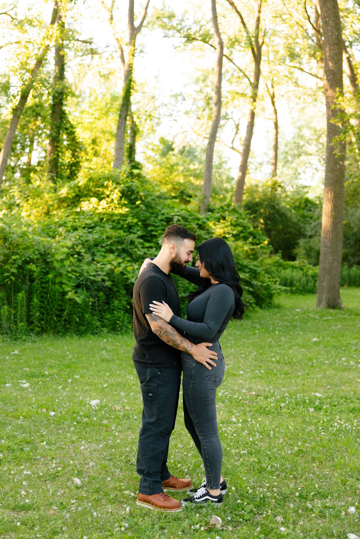 A romantic couple embraces in a sunlit forest clearing wearing matching dark clothing.