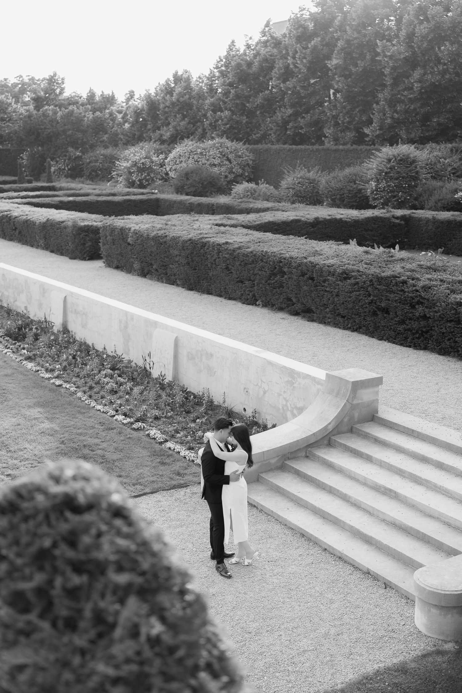 A black and white capture of an intimate moment on stone steps amidst perfectly manicured garden grounds.