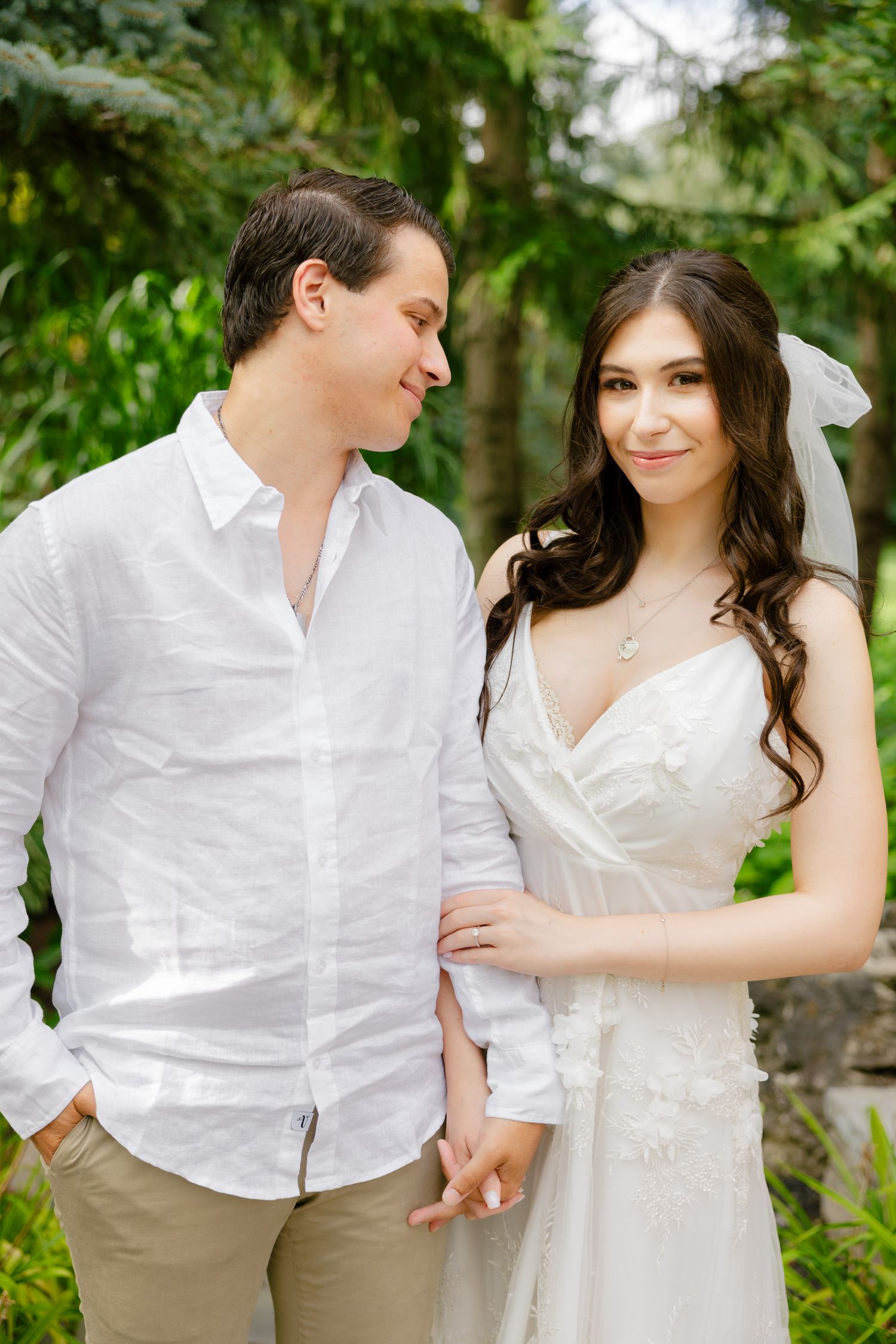 A tender moment between newlyweds as they pose in a garden with evergreen trees in the background.