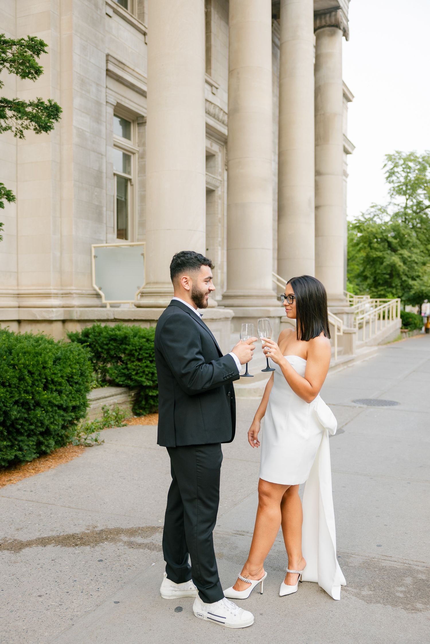 Couple in formal attire sharing champagne toast outside classical building.