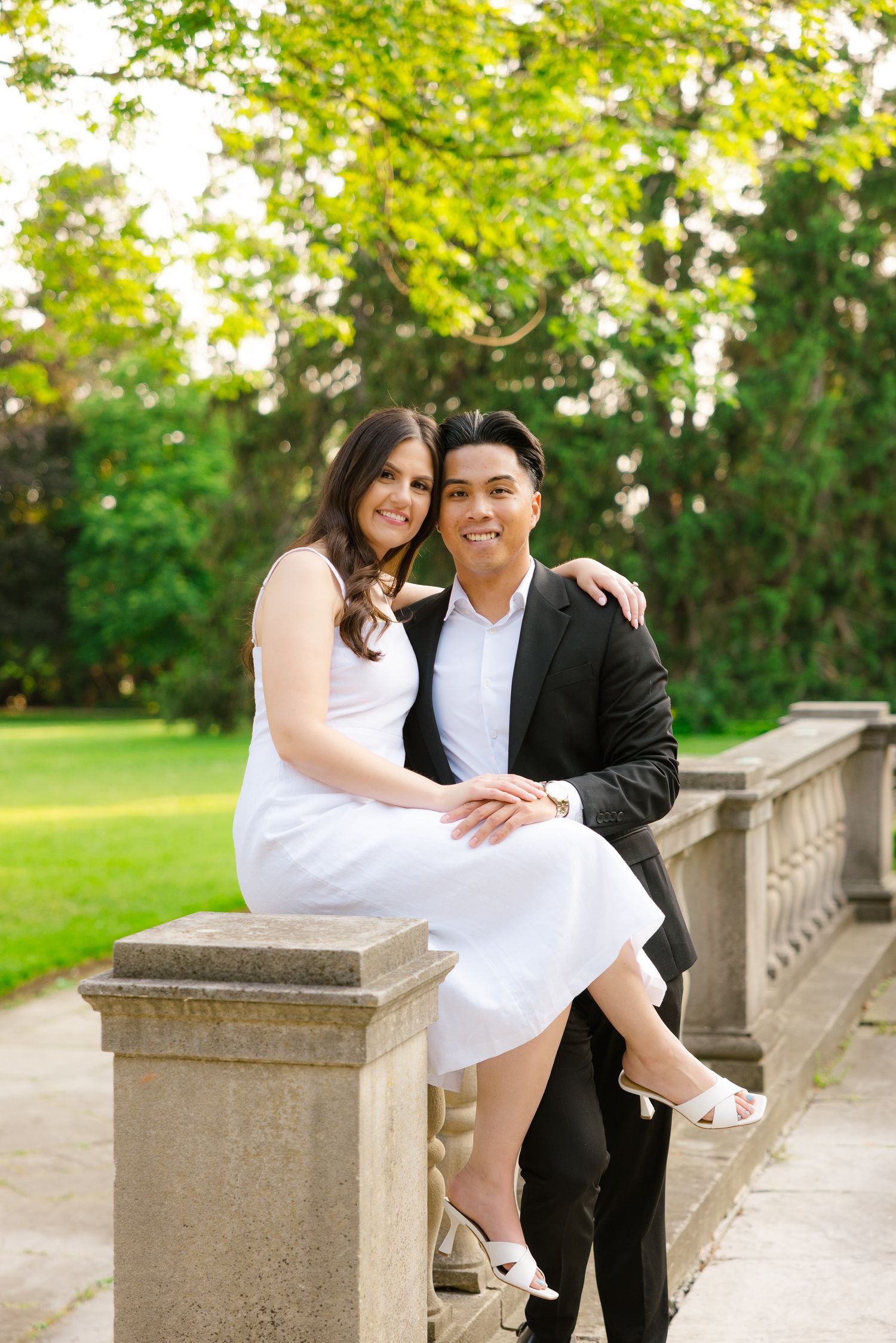 Romantic couple sitting on stone railing in garden with green trees and foliage in background.