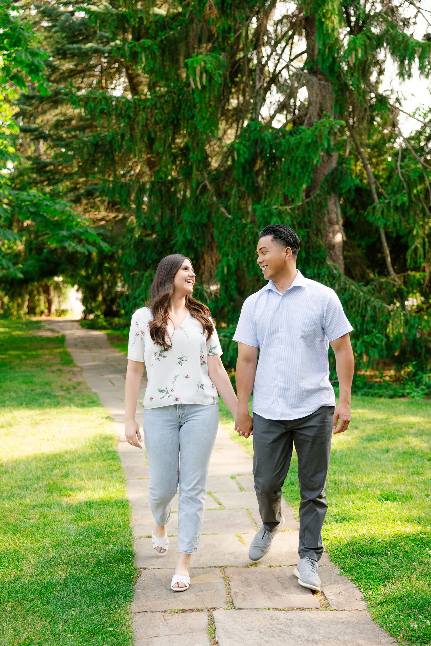 A couple walks together on a stone path through a garden with tall evergreen trees in the background.