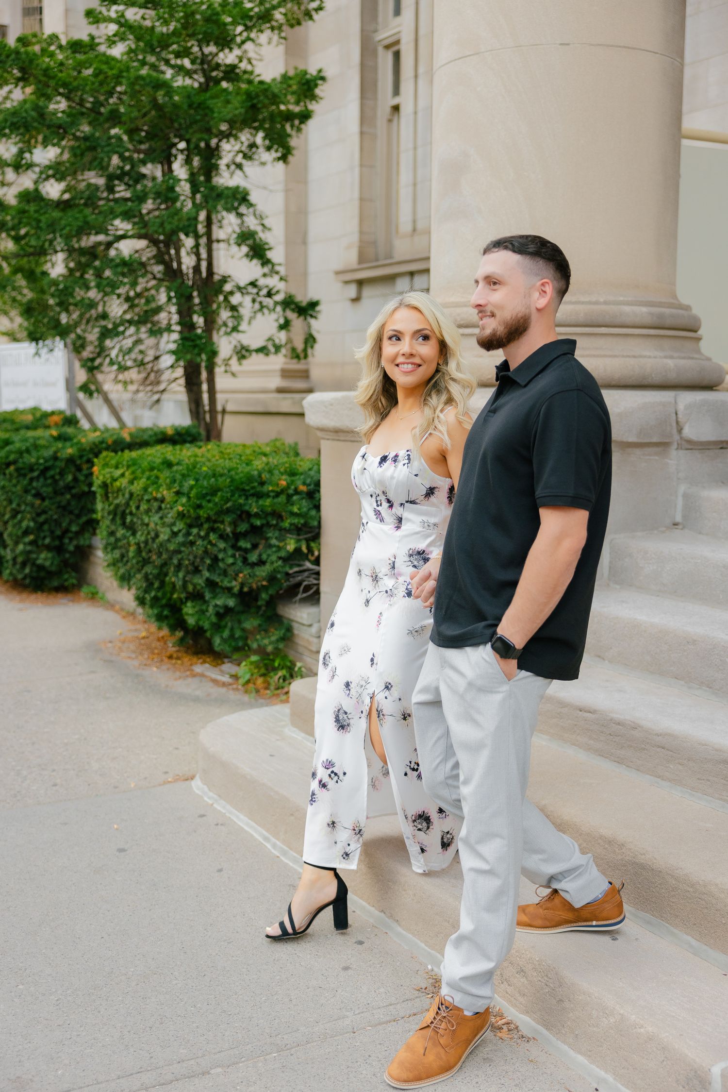 A couple stands by ornate stone columns and greenery outside a historic building entrance.
