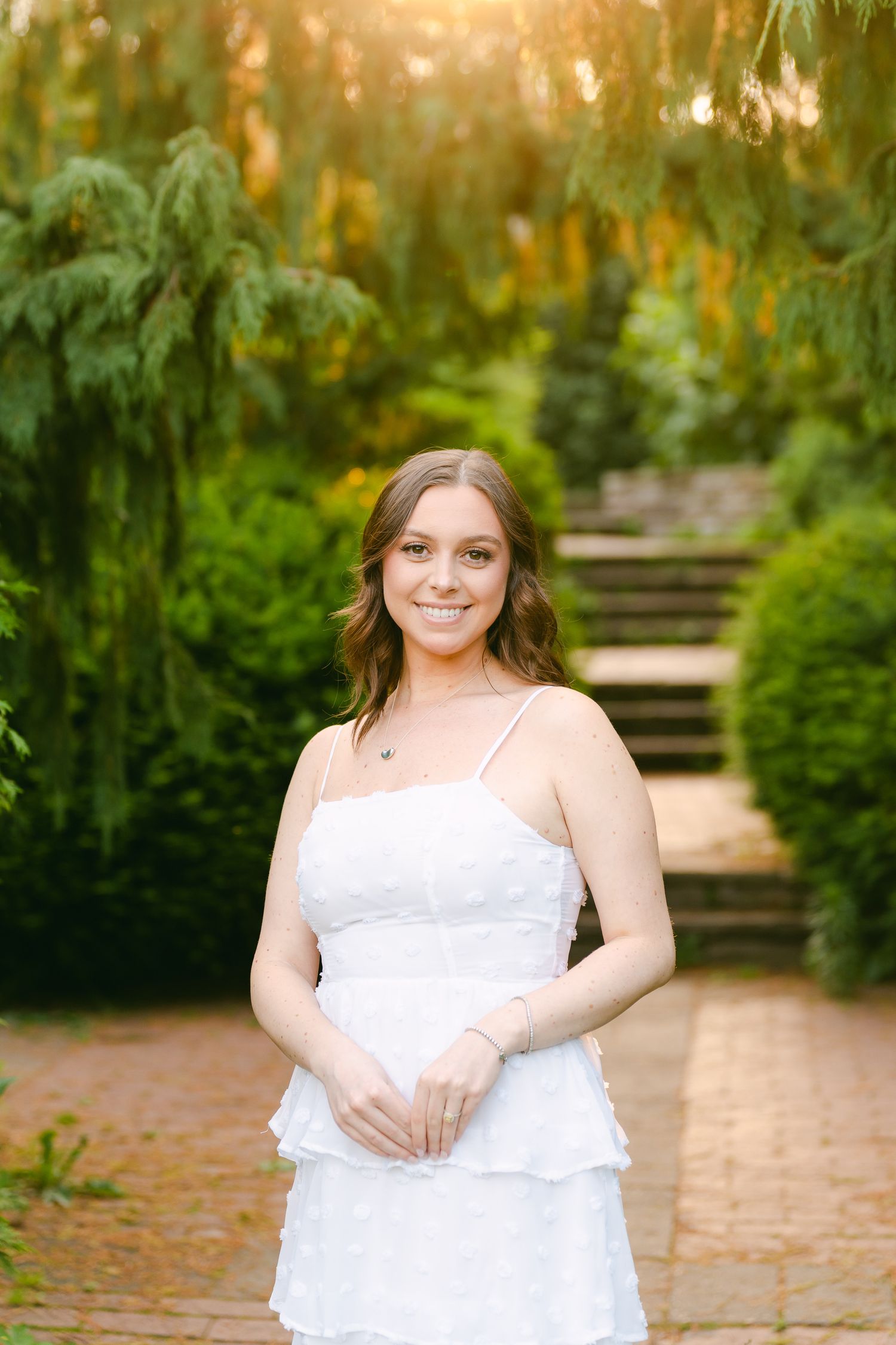 Woman in white summer dress smiles at camera while standing in a garden pathway.