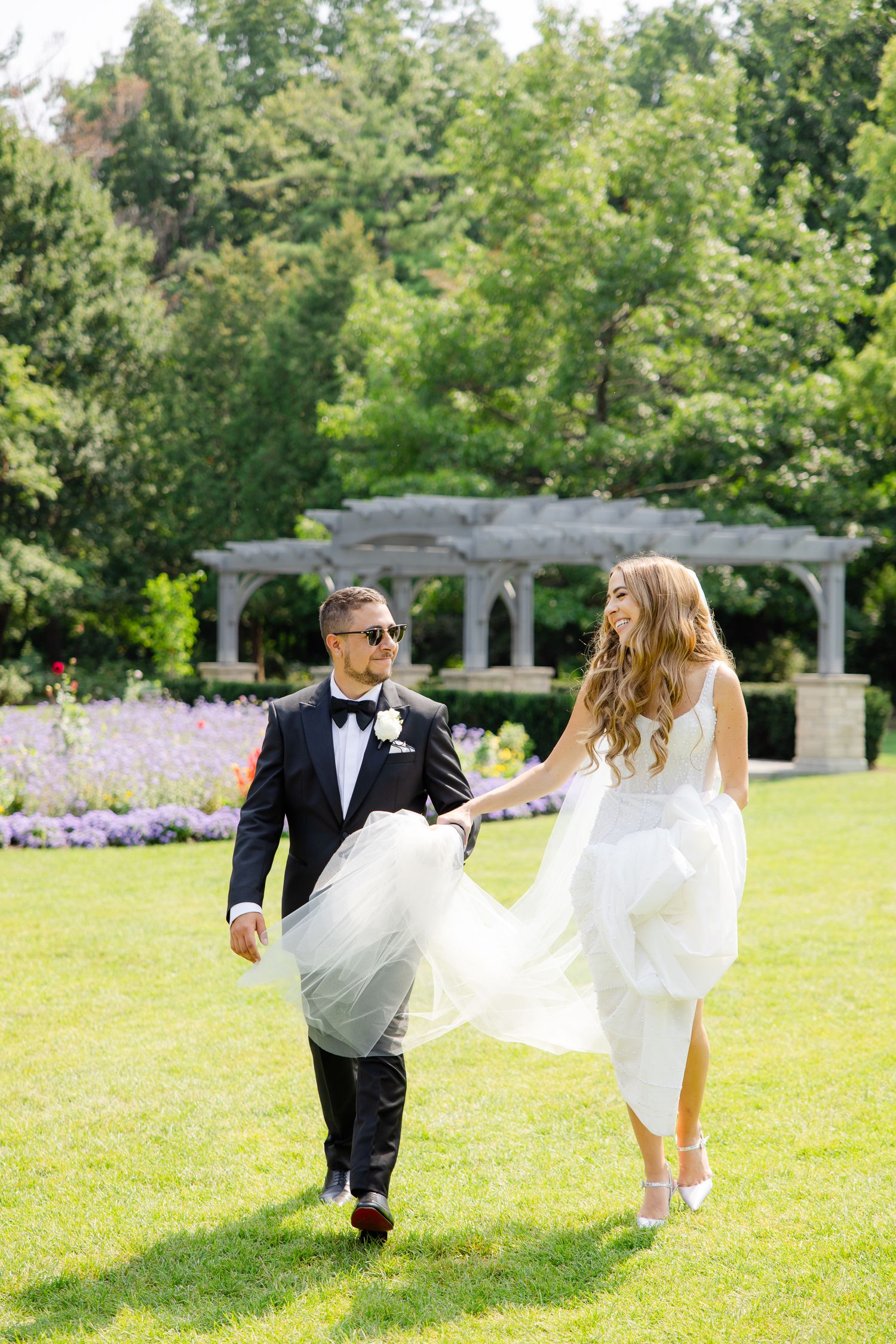 Wedding couple strolls through a sunlit garden with a white pergola and purple flowers in background.