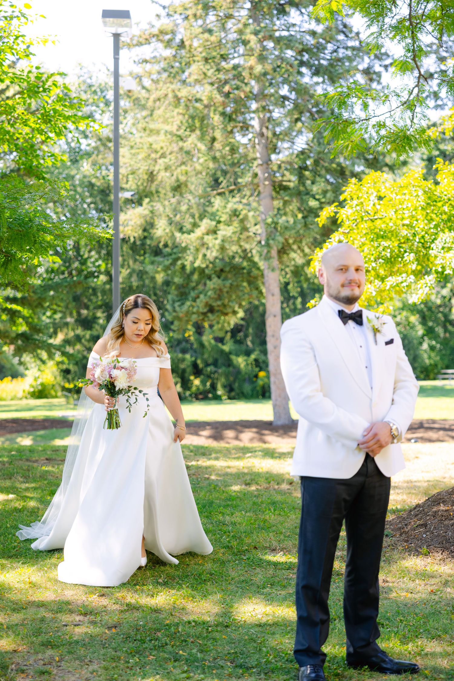 First look wedding photo in sunny garden with white tuxedo and flowing bridal gown.