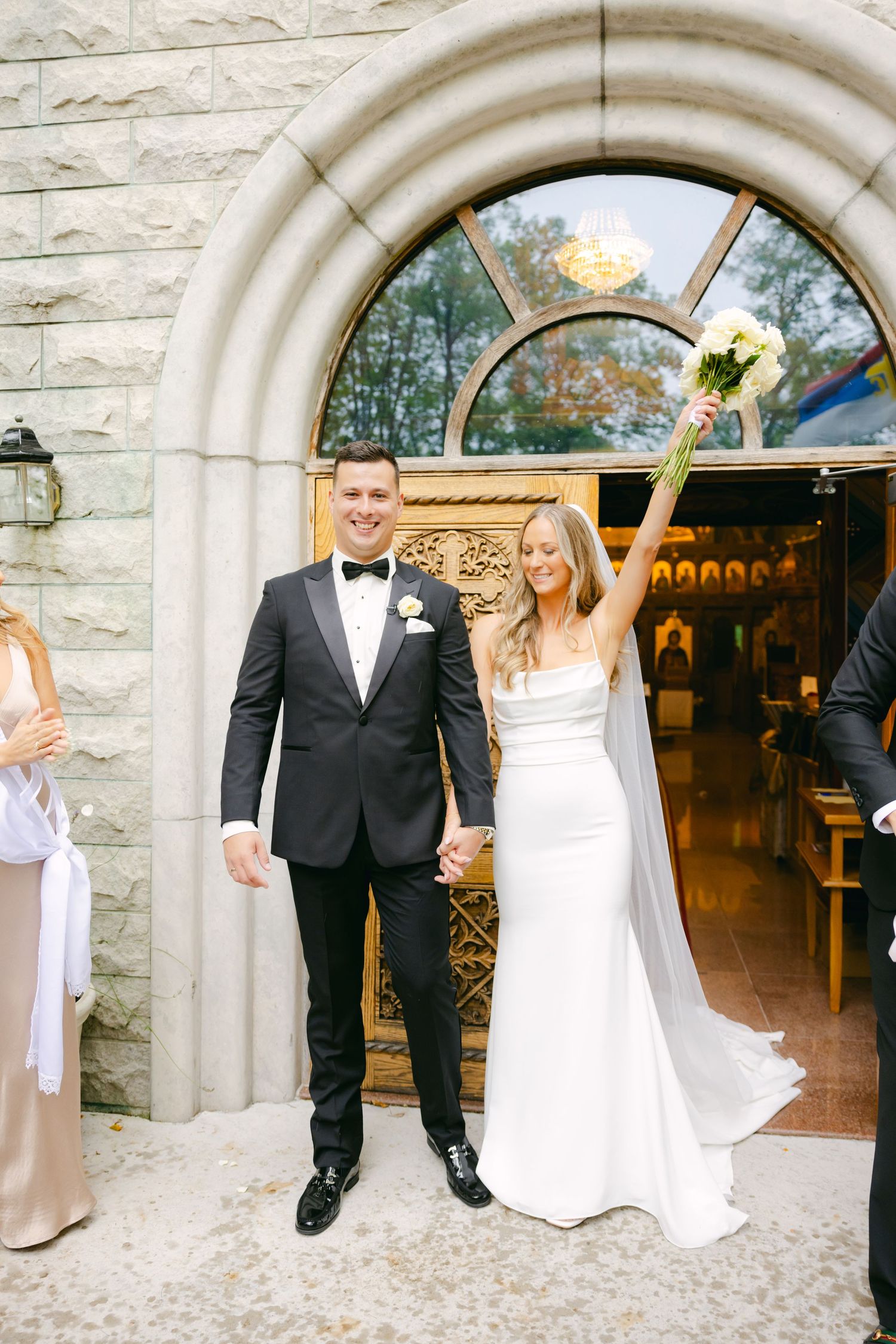 A wedding couple stands in front of an elegant stone archway entrance at a church.