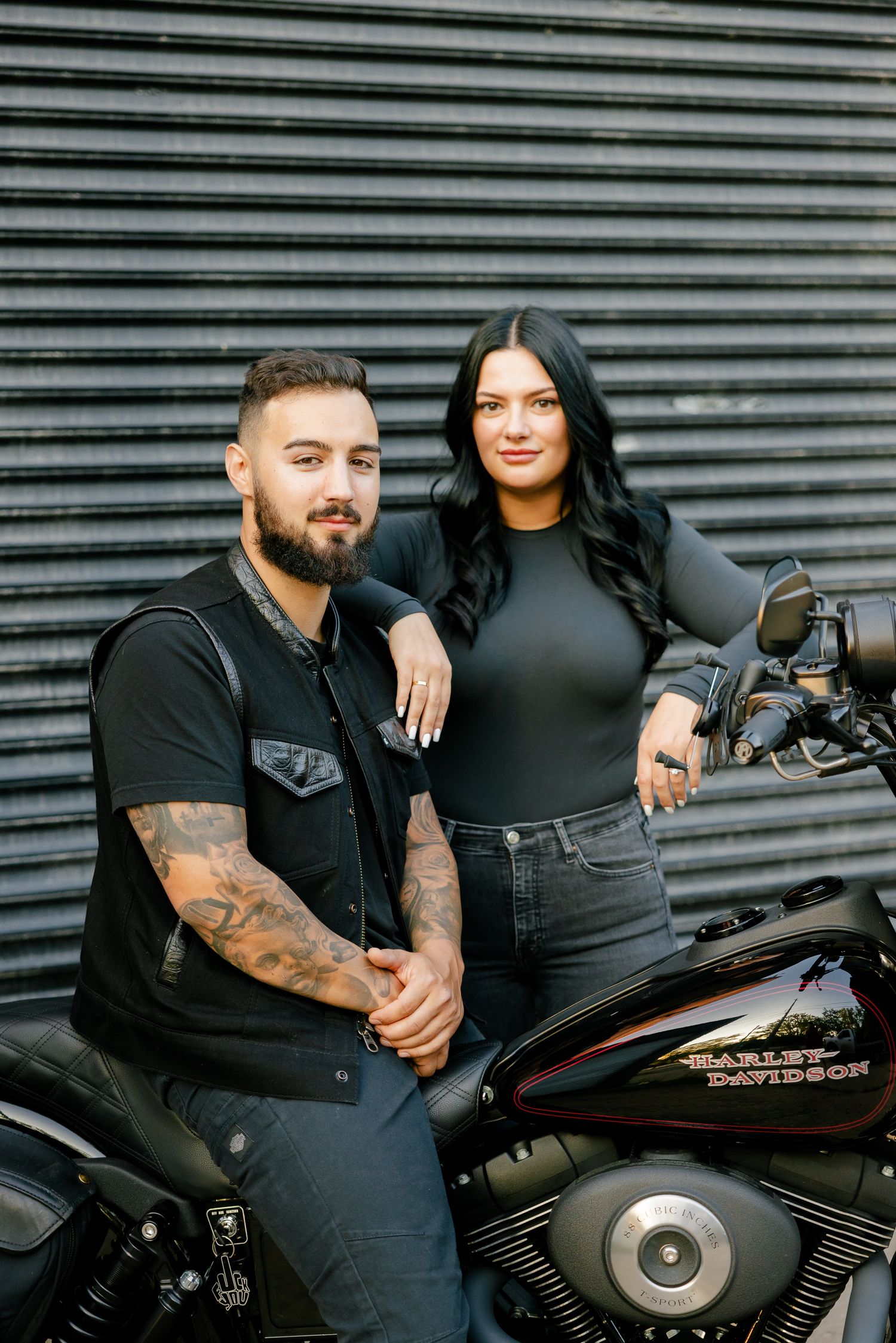 A couple stands beside their motorcycle for a portrait against an industrial backdrop.