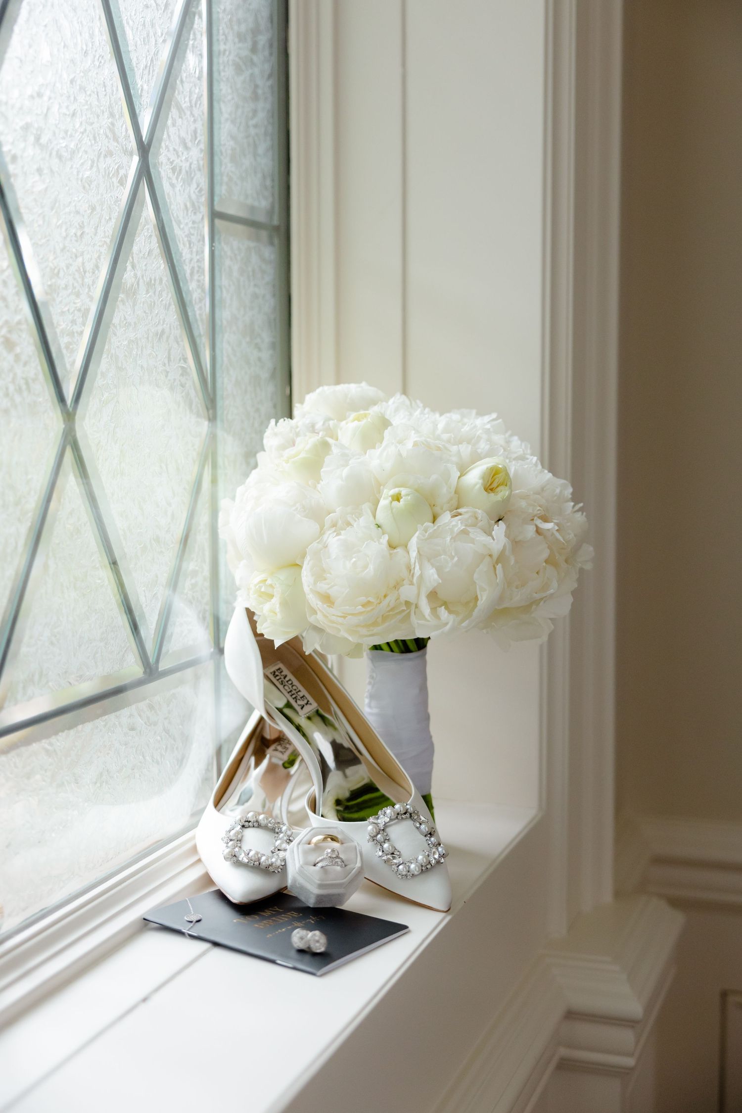 Classic white wedding shoes and peony bouquet arranged on window ledge with decorative glass.