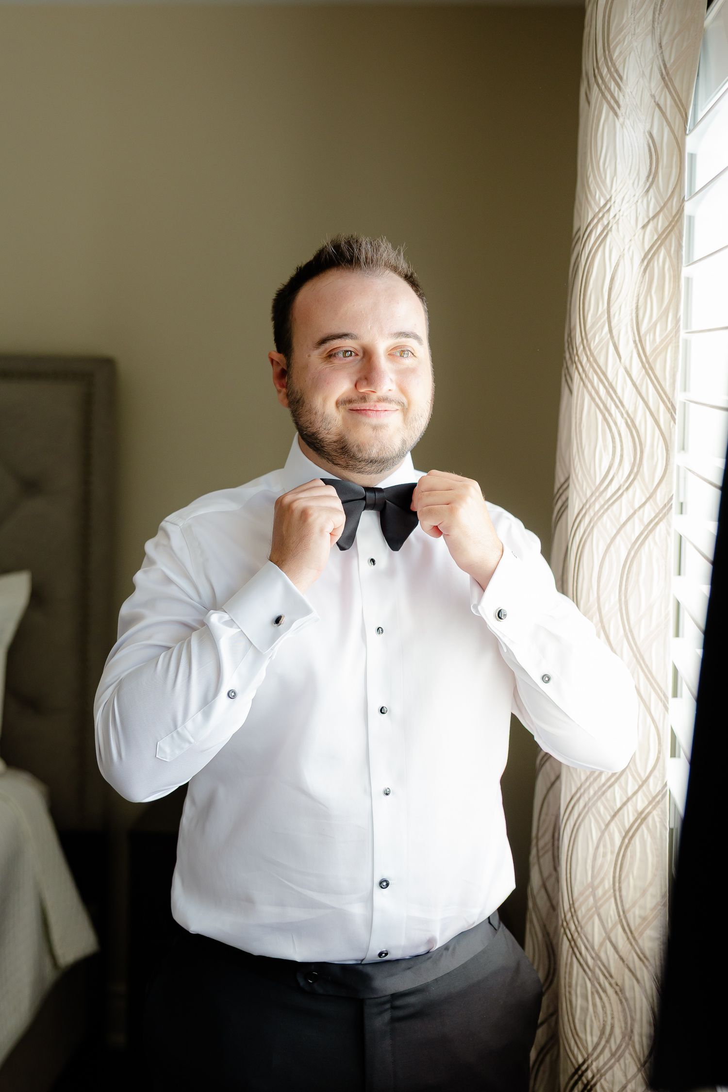 Man adjusts his white bowtie while wearing a tuxedo with black vest.