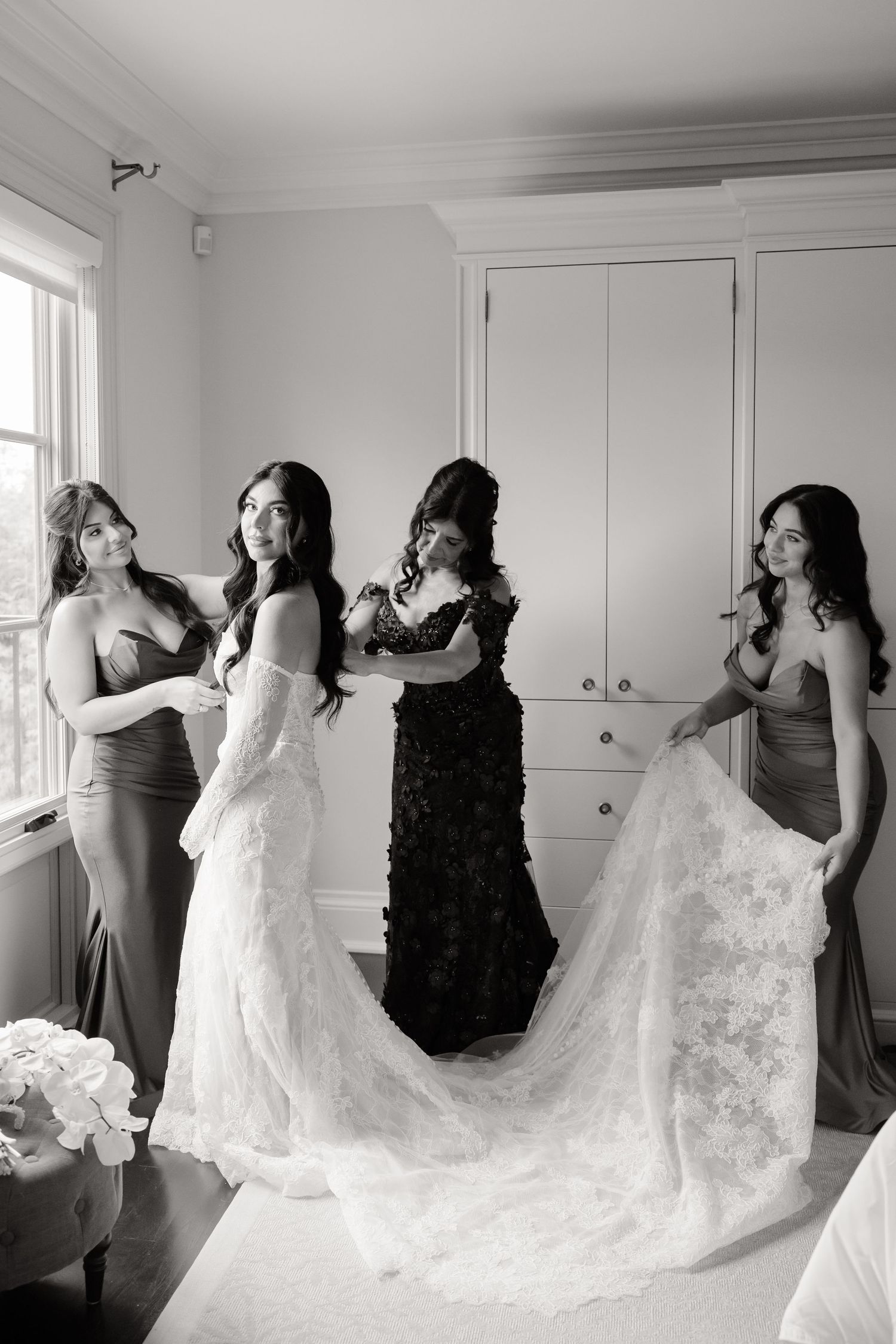 A group of women in formal dresses help prepare and adjust a white wedding gown with a flowing train in a bright room.