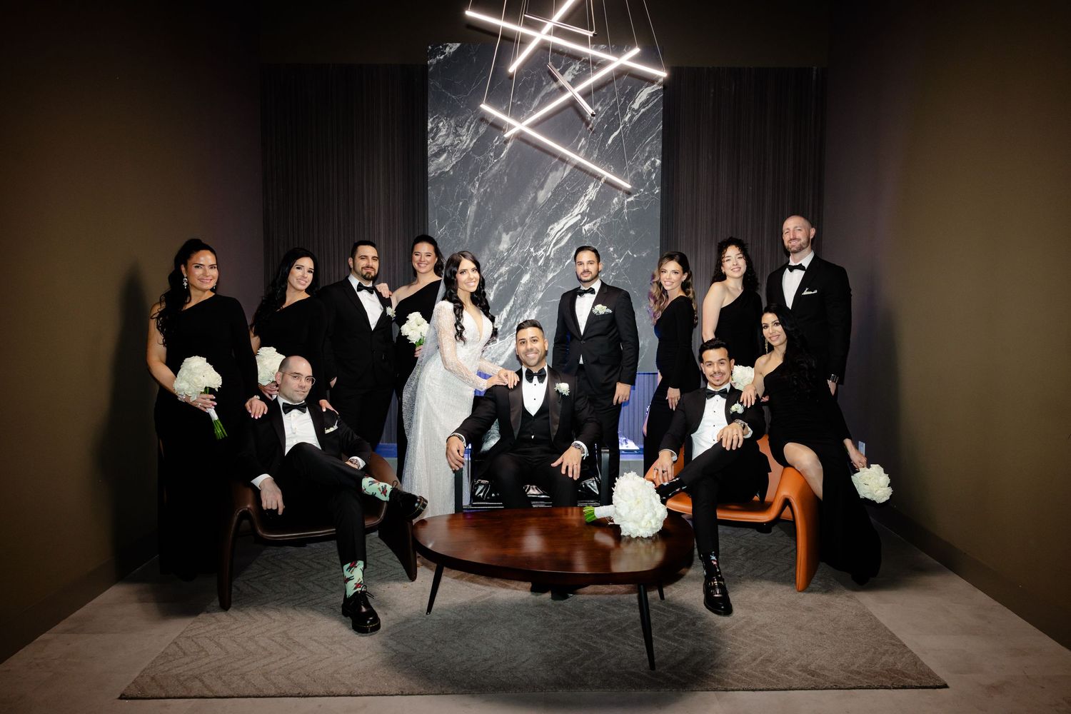 Wedding group portrait features black-clad attendants arranged around modern furniture.