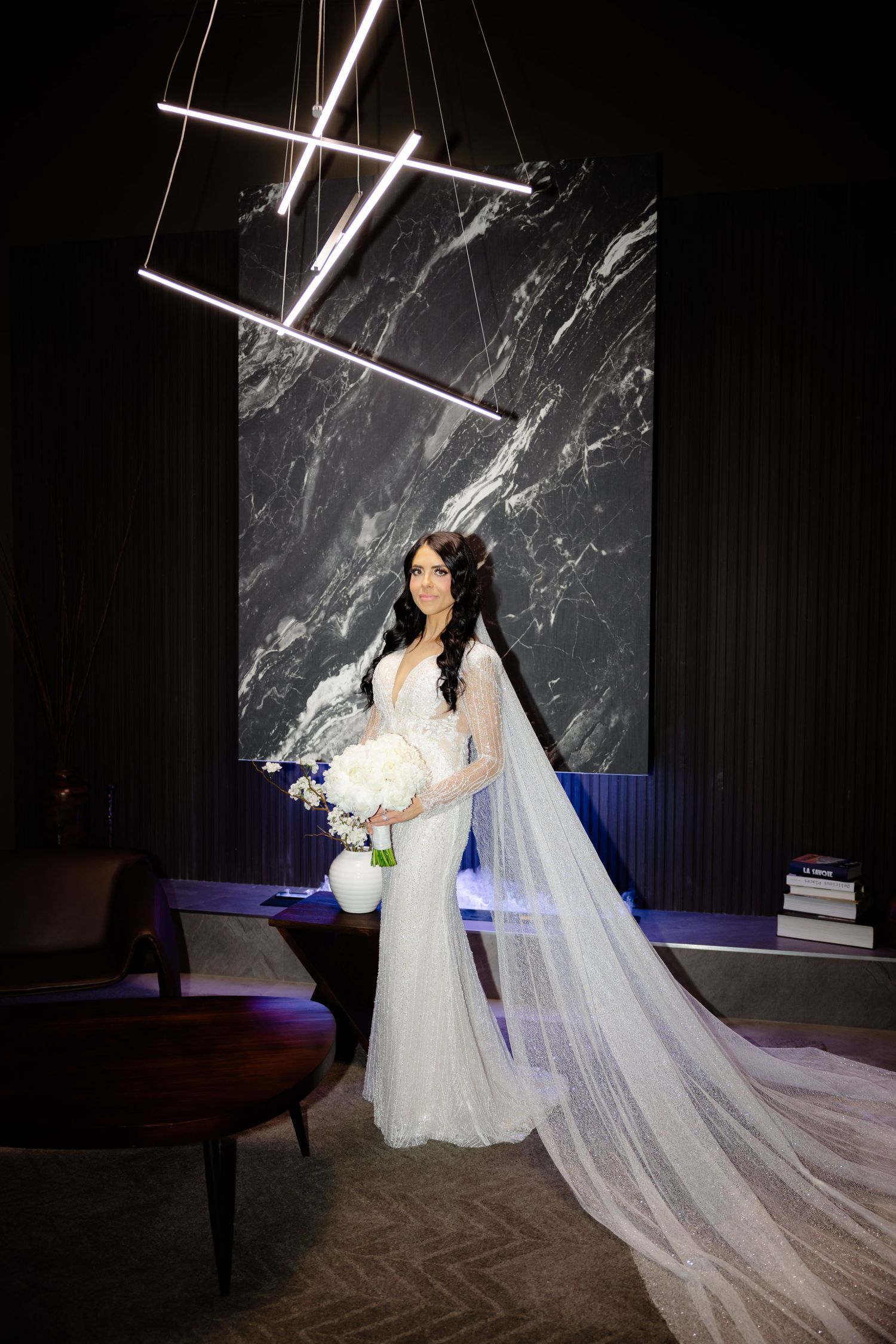 Bride holding white bouquet poses in front of marble wall with geometric light installation.