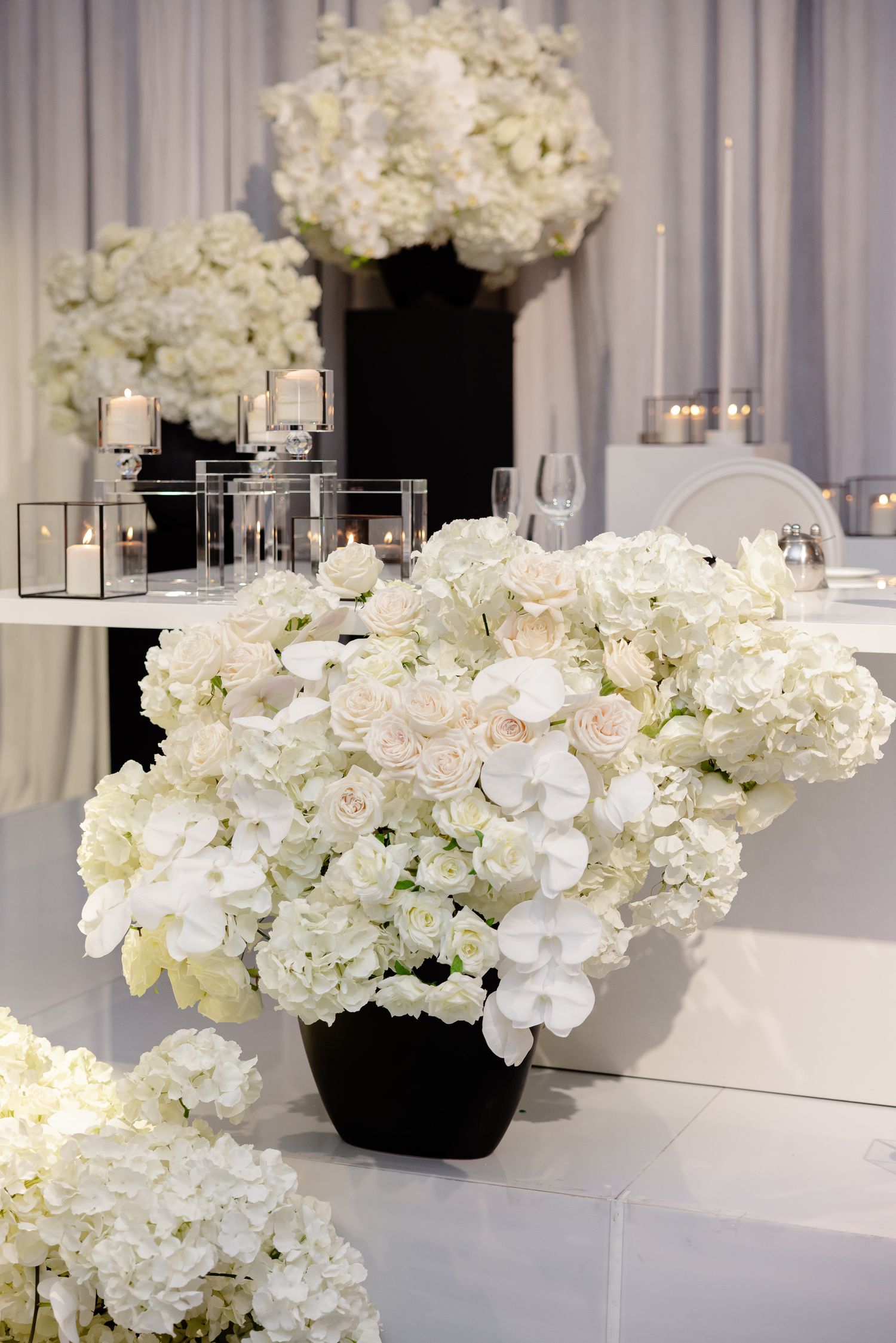 Elegant white hydrangea and orchid floral arrangements in black vases with modern candle holders on white table setting.