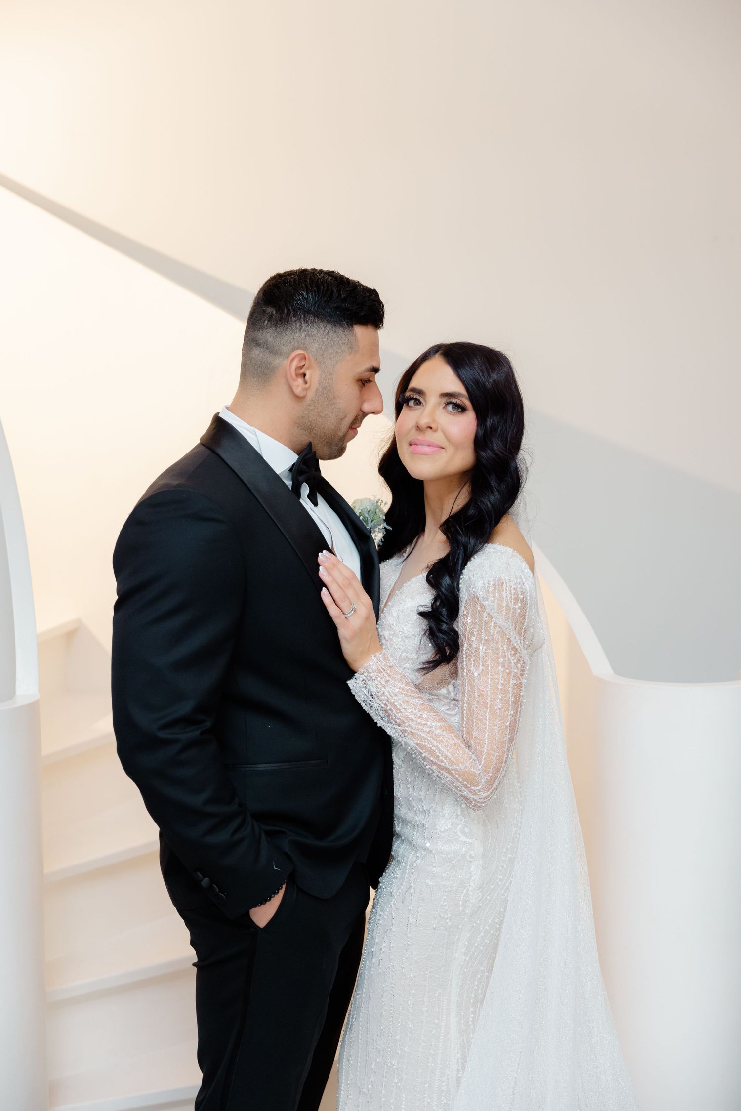 A romantic wedding portrait of a couple in a minimalist white setting, with the bride in lace and groom in black tuxedo.
