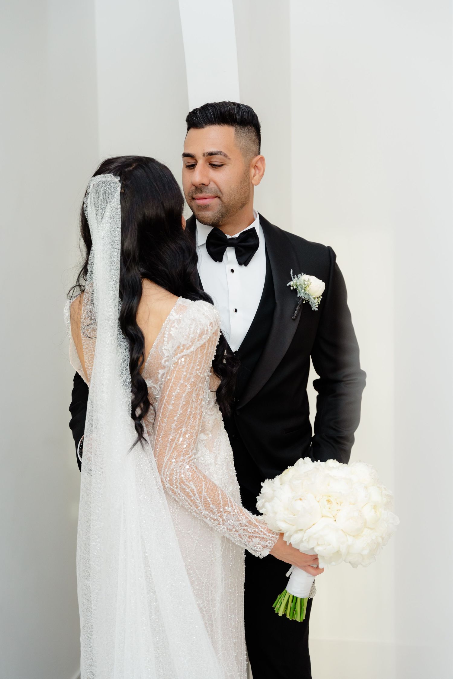 Elegant couple in wedding attire pose intimately against a white wall.