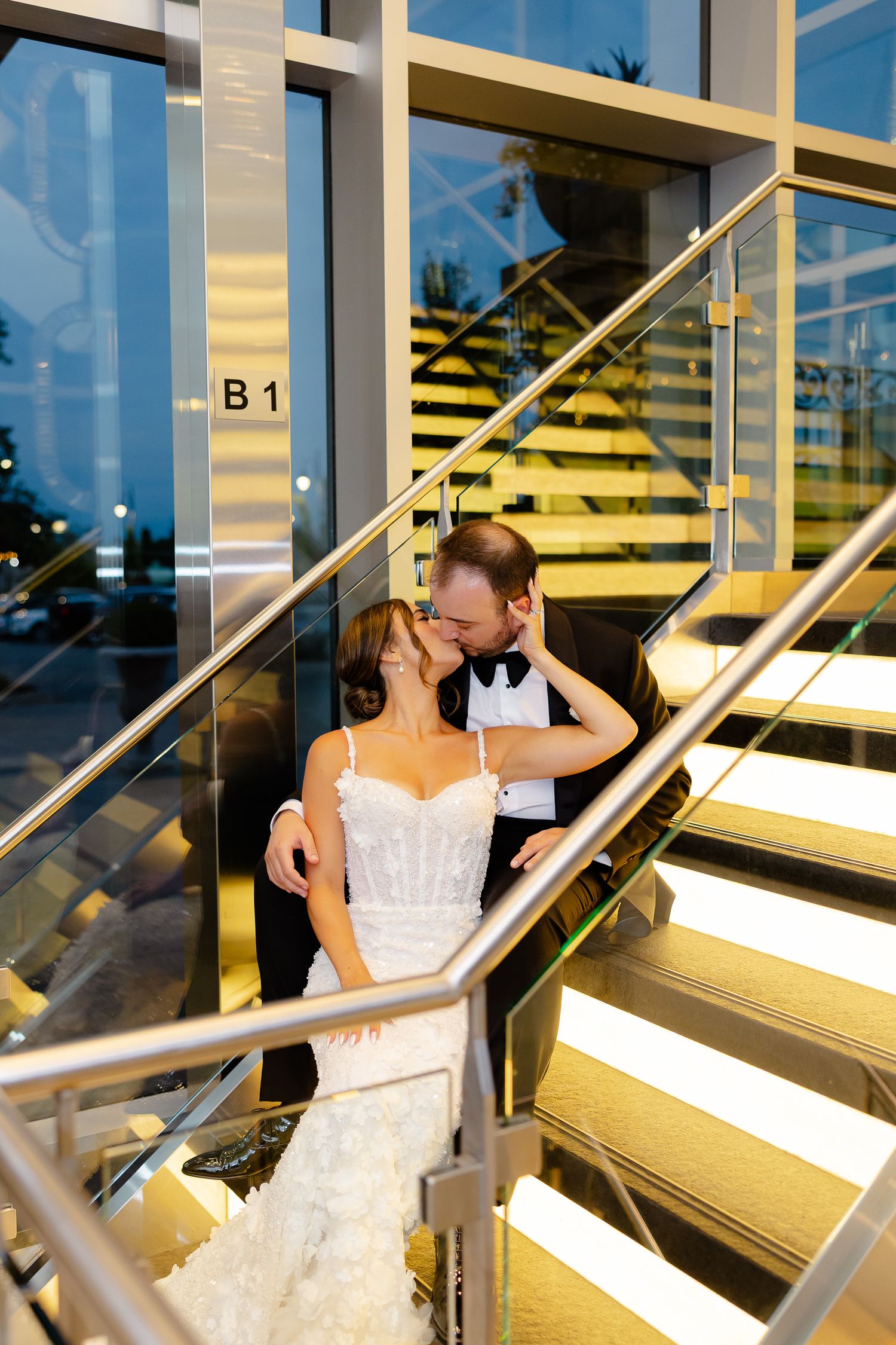 A couple shares a romantic kiss on a modern staircase with glass panels and metal railings during a wedding photo shoot.