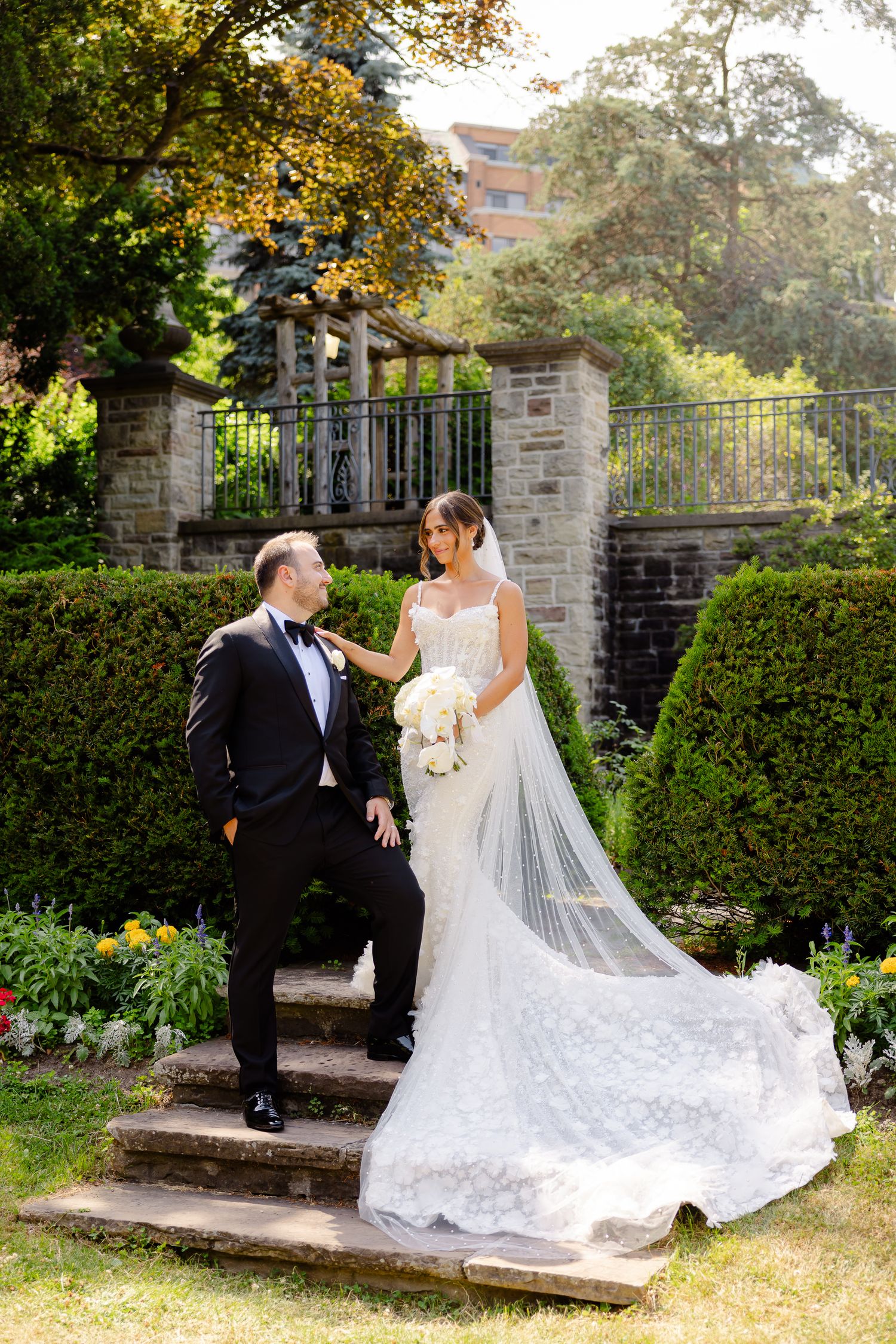 A couple in wedding attire stand together on stone steps in a lush garden setting with ornate iron gates.