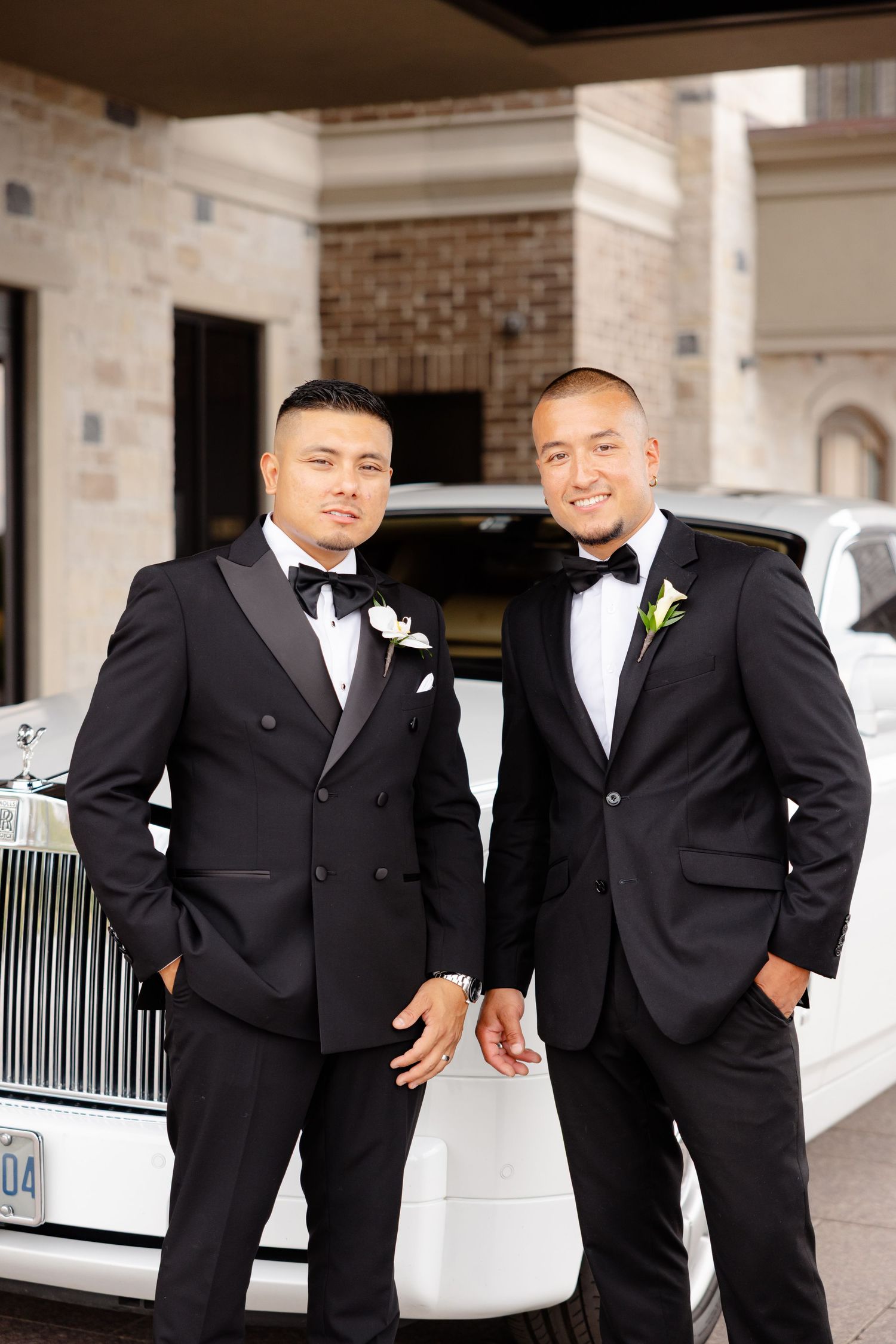 Two groomsmen in black tuxedos and bowties stand next to white luxury vehicle at elegant venue entrance.