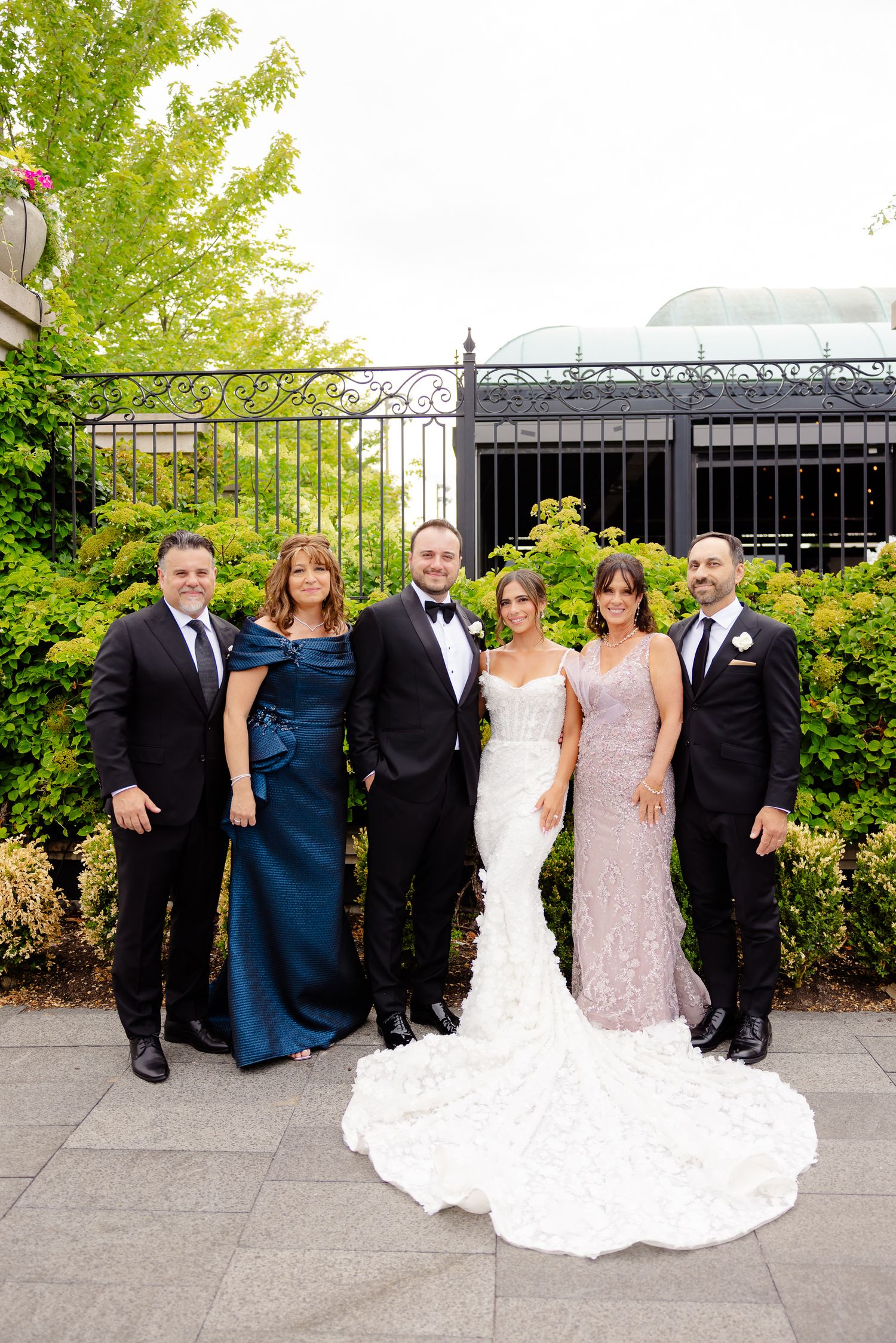 Group in formal attire gathered at an outdoor garden venue with decorative iron gates and greenery in the background.