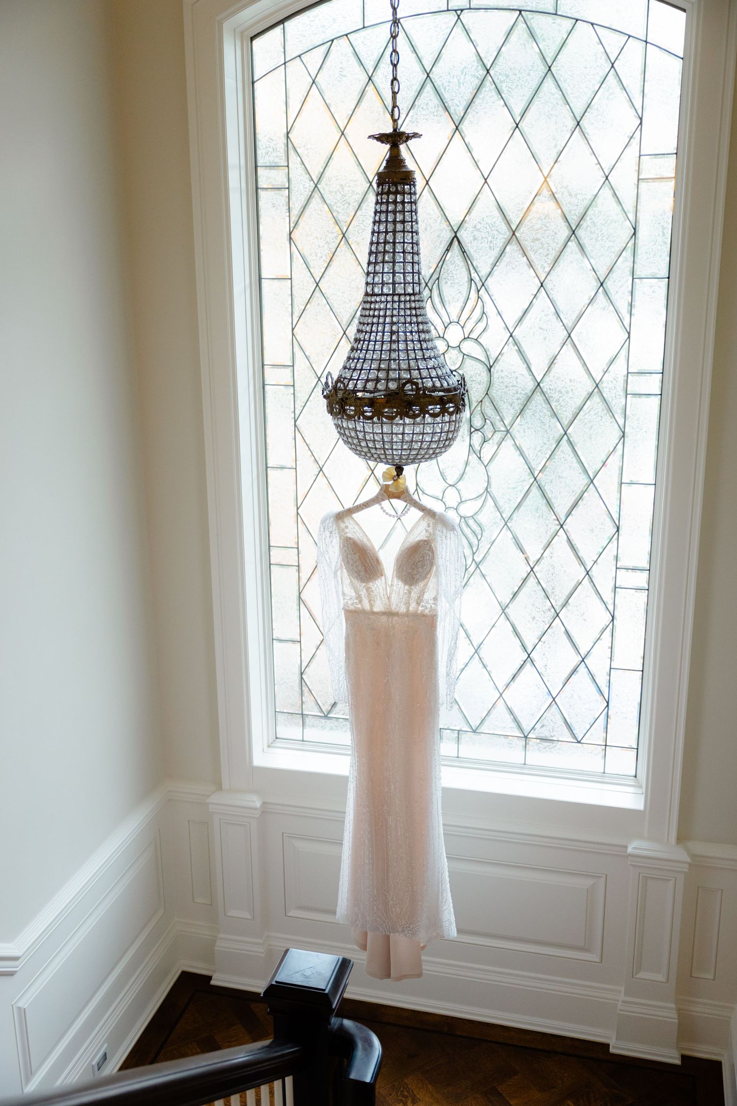 Long champagne wedding gown displayed under crystal chandelier in front of arched window.