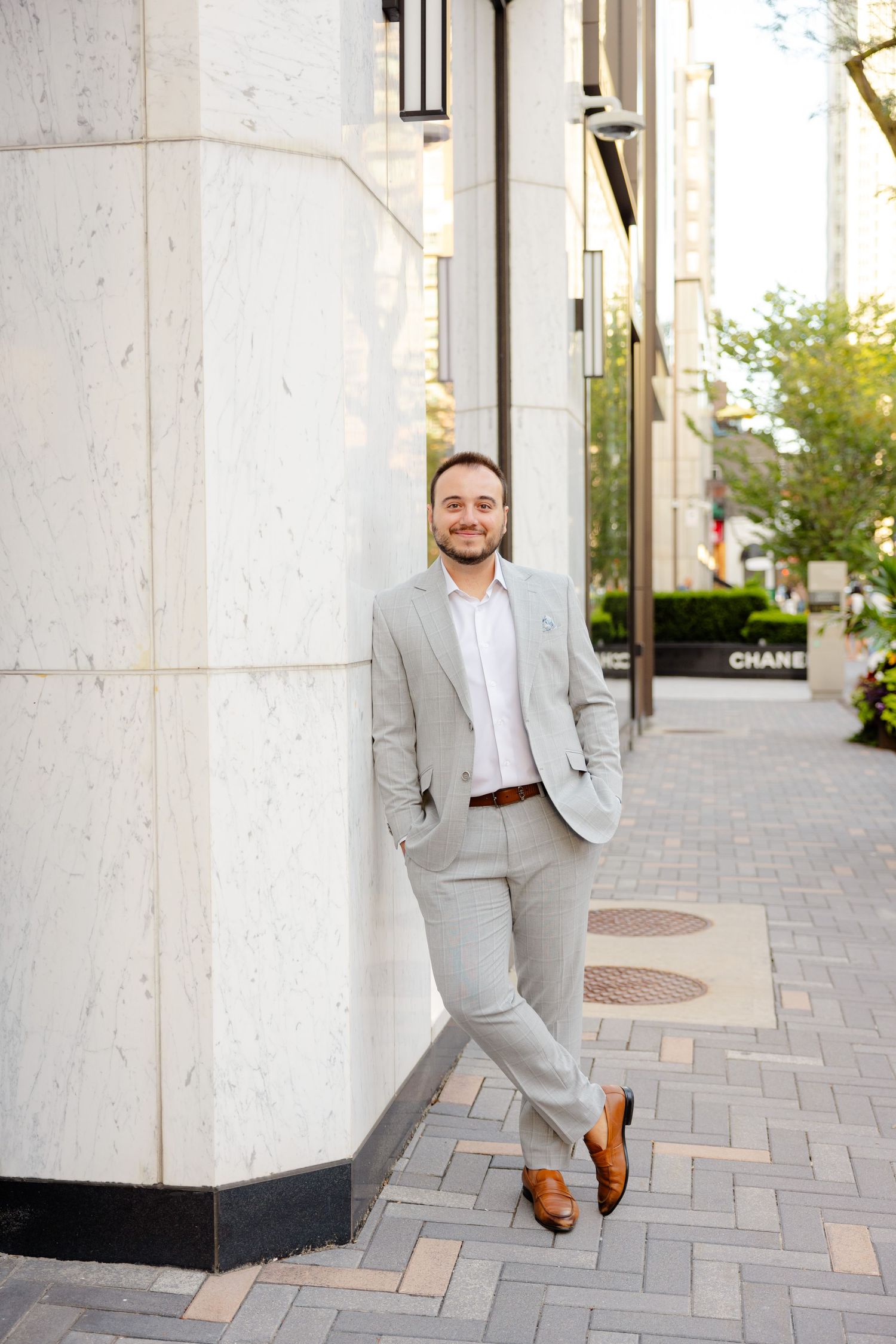 A stylish man in a light gray suit leans against a white building column on a brick sidewalk.