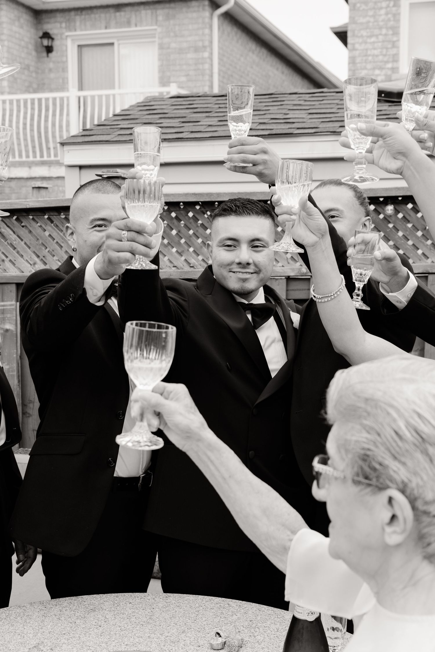 Groomsmen in formal attire raise glasses in a celebratory toast at an outdoor wedding reception.