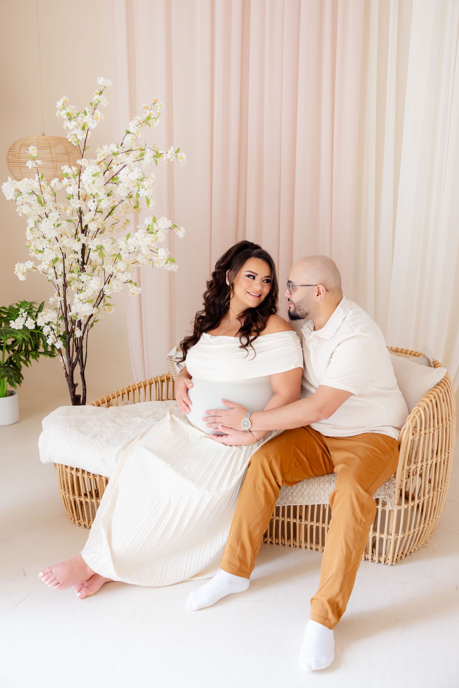 A couple sits together on a rattan loveseat with white flowers and pink curtains in the background.