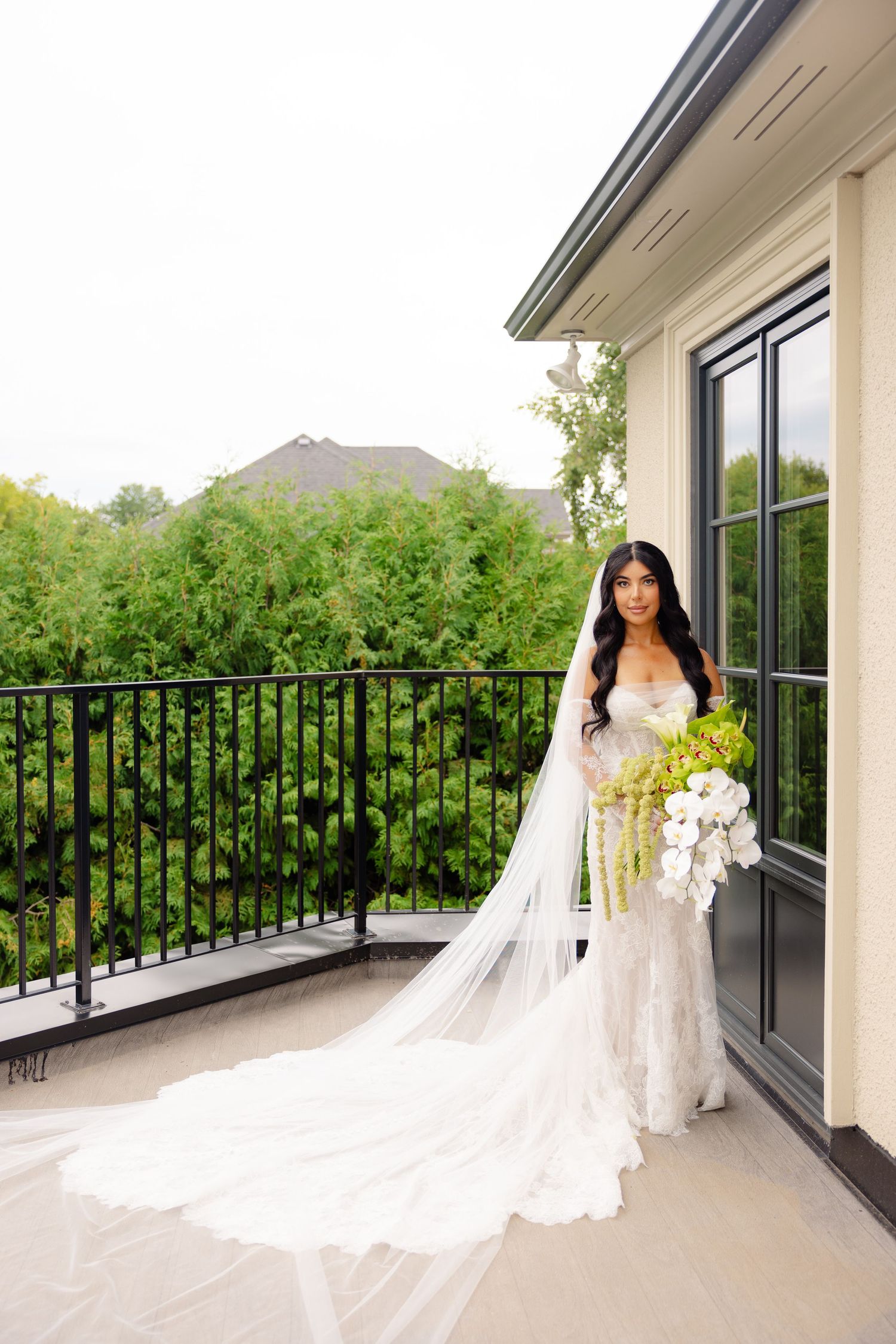 A stunning bride in a flowing white wedding gown with long veil stands elegantly on a covered balcony.