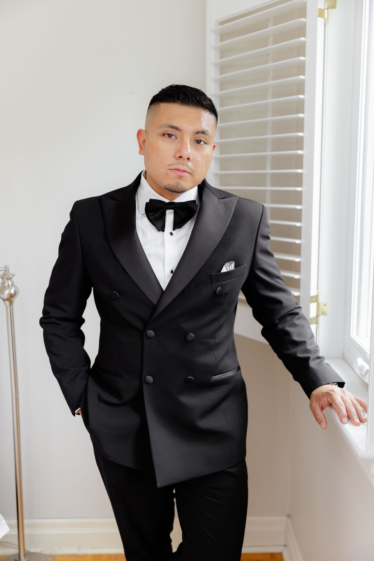 Well-dressed person in a black double-breasted tuxedo with white dress shirt and bow tie leaning against window.