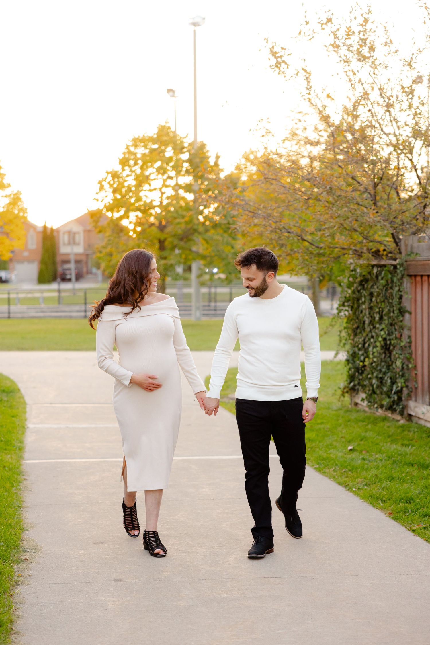 A couple in white outfits walks hand in hand on a sidewalk with autumn trees in the background.