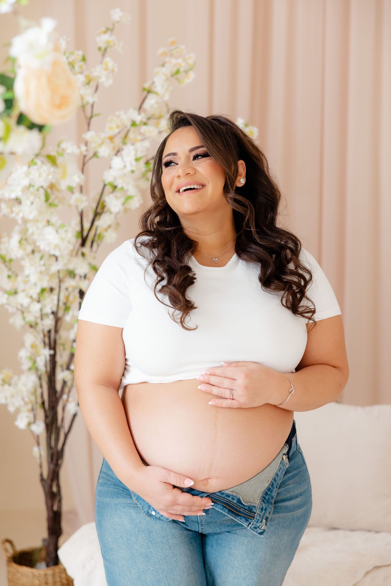 Expecting mother in white t-shirt and jeans poses with flowers while holding her belly and smiling warmly.