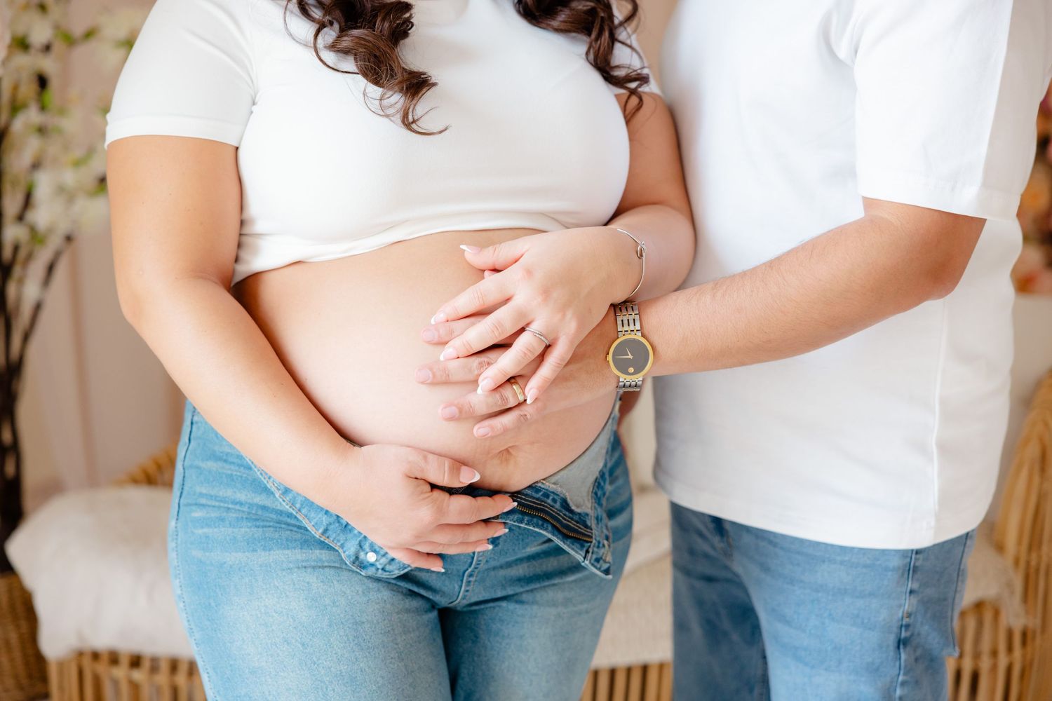 A couple in white shirts and jeans embrace while holding a pregnant belly in a tender maternity moment.