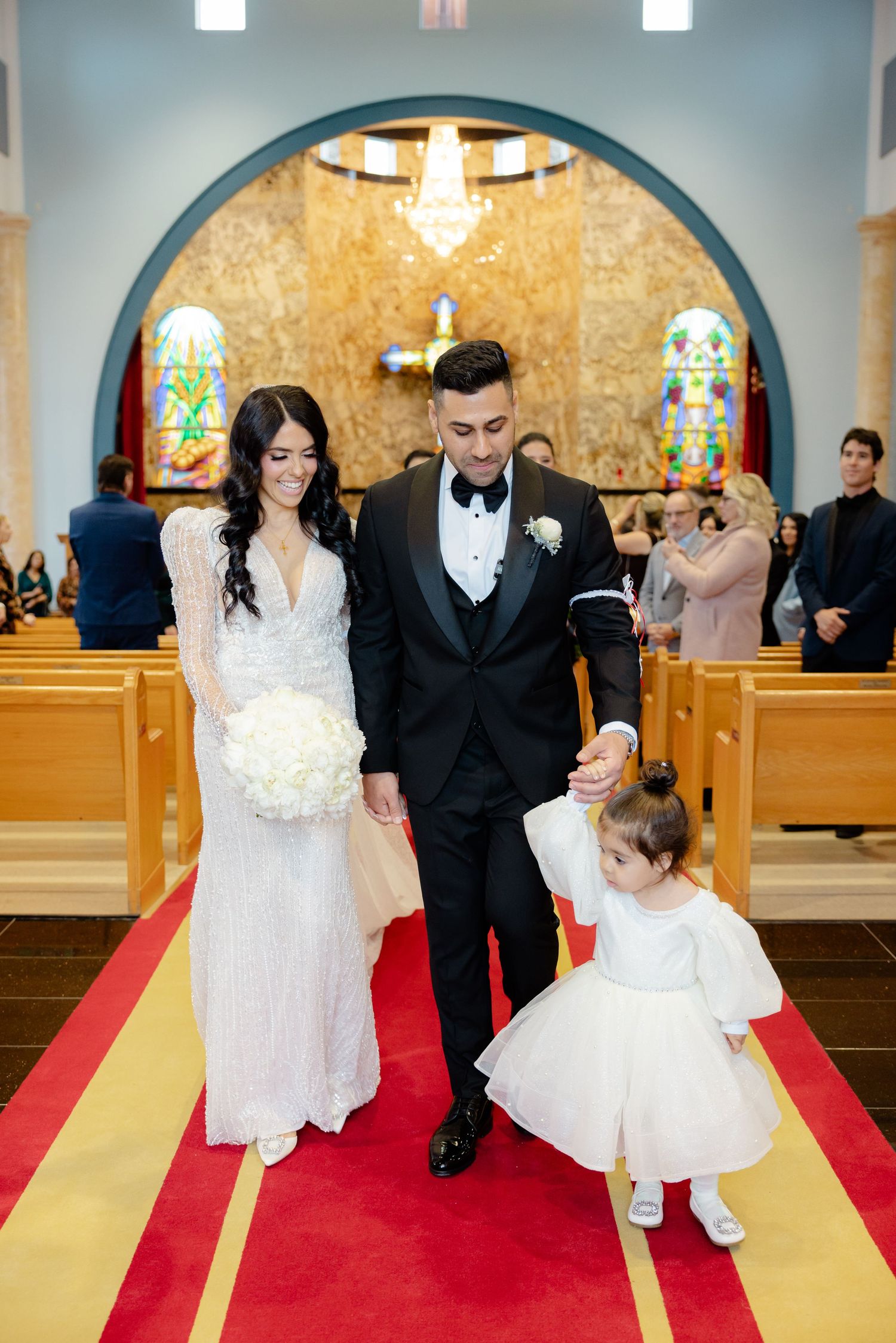 Wedding party exits down red carpeted church aisle beneath arched ceiling and stained glass.