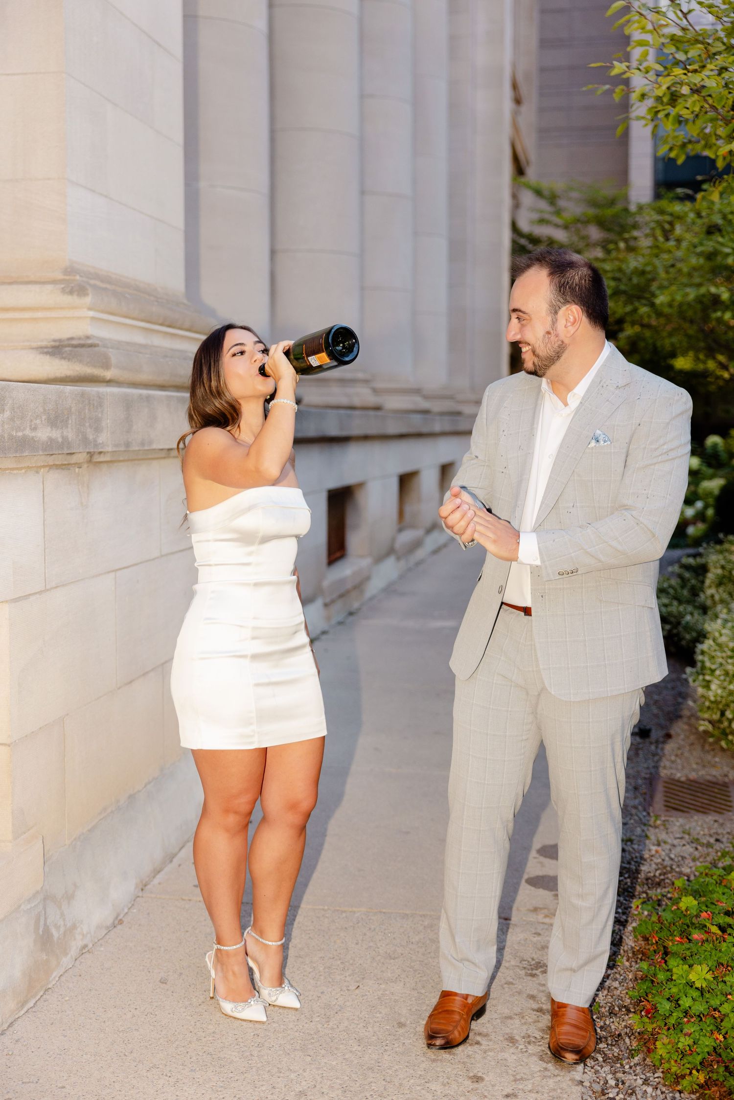 A woman takes a sip from champagne while her companion watches by elegant stone columns.