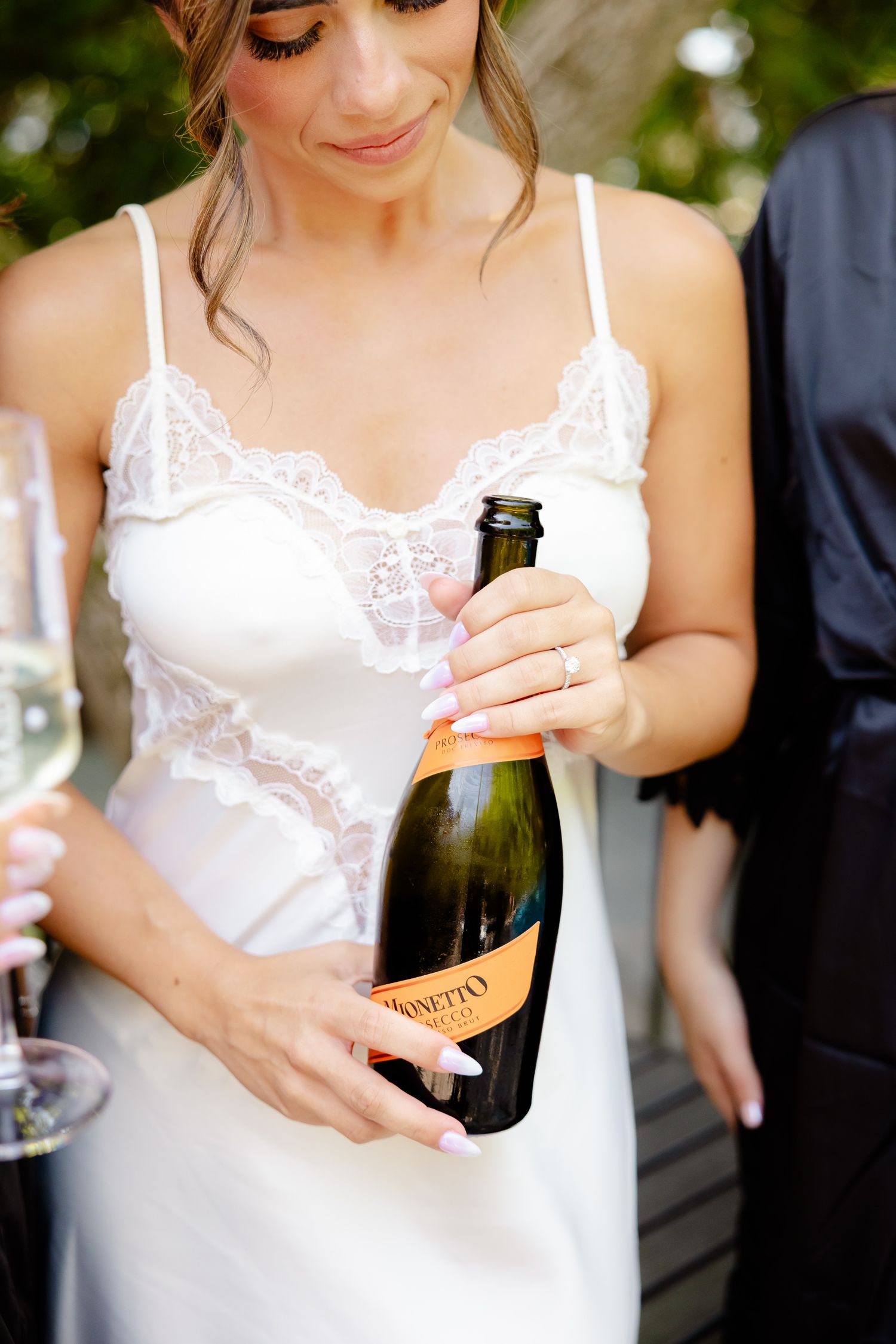 Series of close-up shots showing hands in white lace garments holding and opening champagne bottles outdoors.