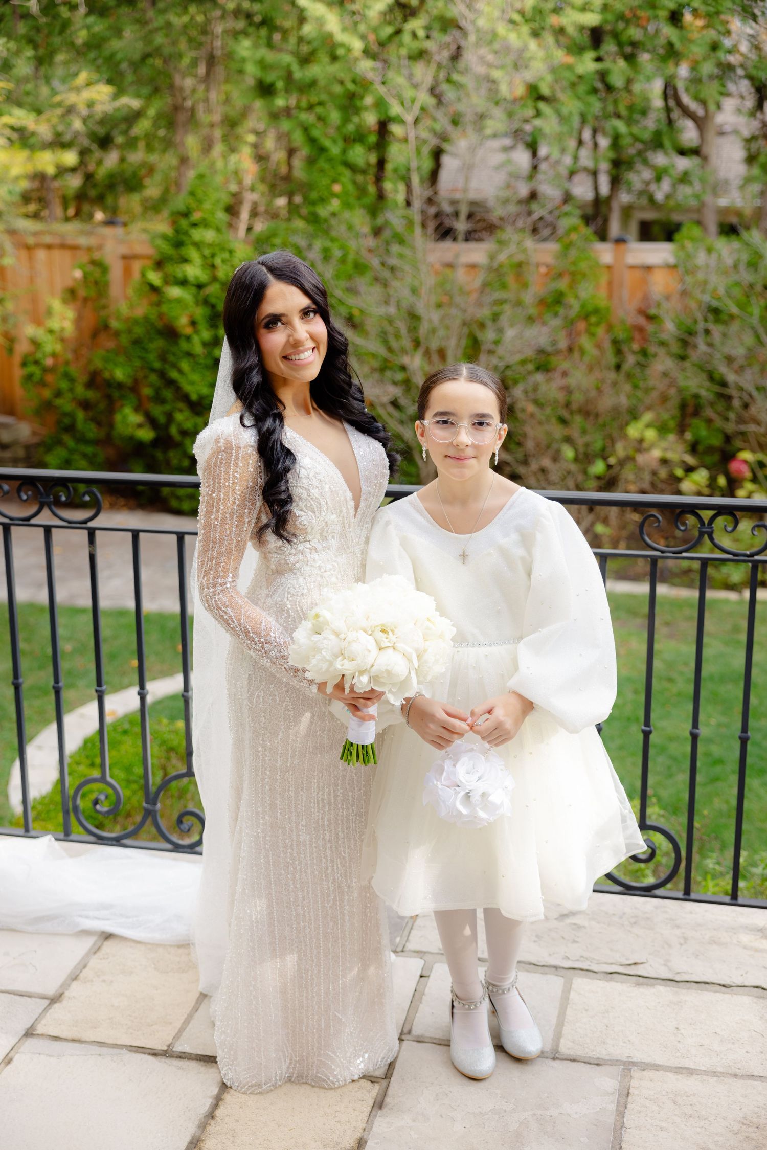 Bride in beaded gown stands with flower girl in white dress holding bouquets on stone patio.