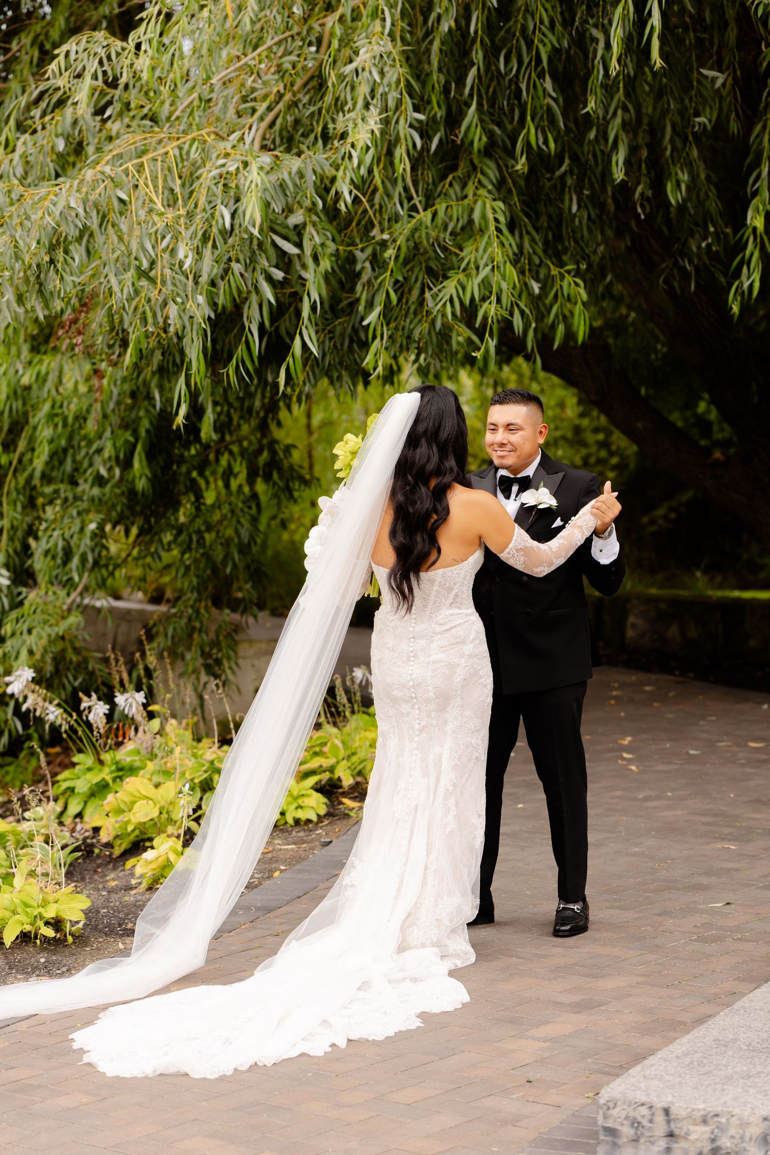Wedding couple posing together under willow trees in an outdoor garden setting.