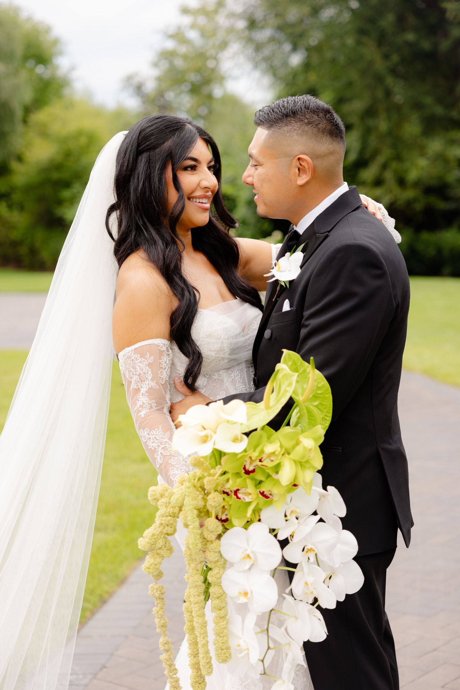Happy newlyweds embrace outdoors on their wedding day with elegant orchid and greenery bouquet in hand.
