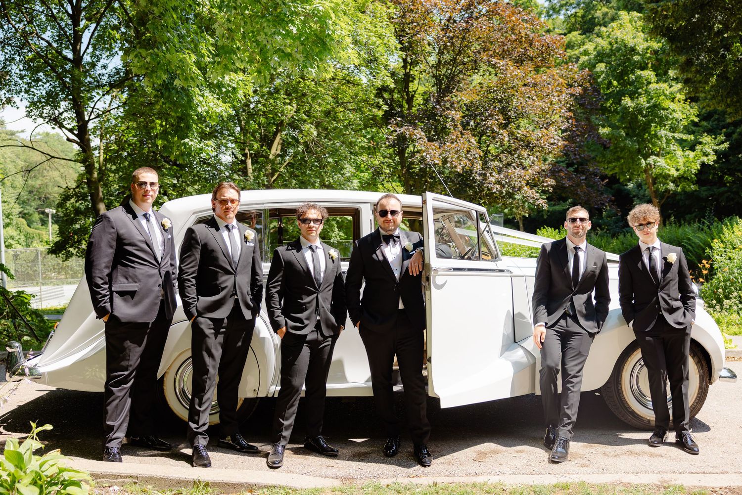 A wedding party in formal black suits pose together outdoors against a backdrop of lush green trees.