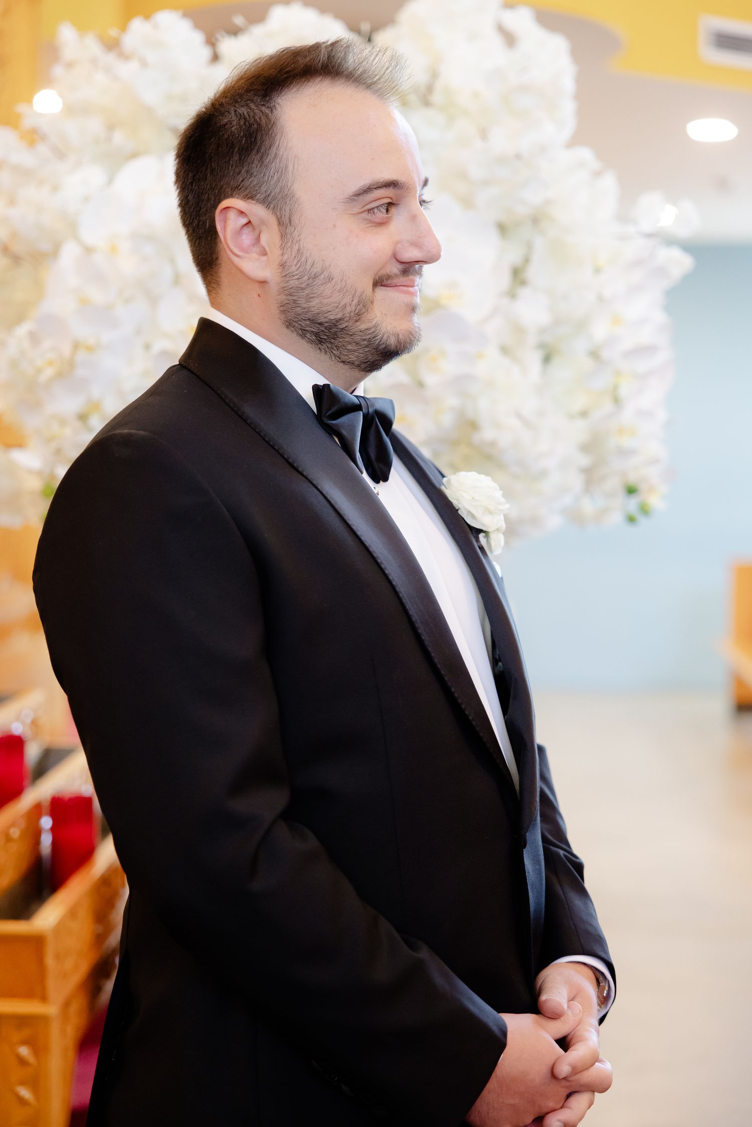 A groom in a classic black tuxedo smiles while standing in front of white cherry blossoms at a wedding ceremony.