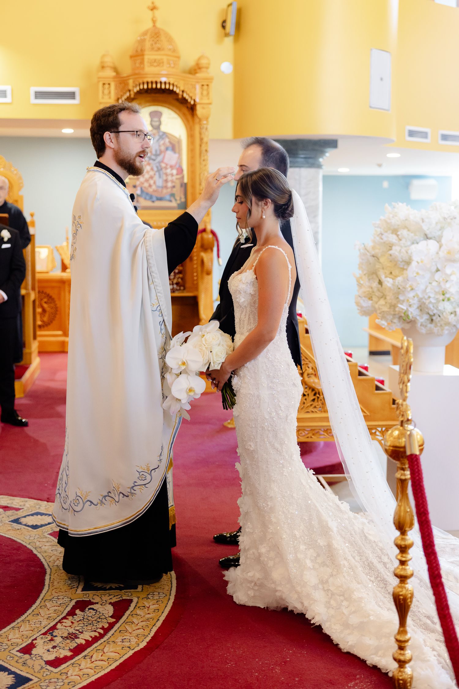 An Orthodox wedding ceremony takes place inside a church with ornate decorations and red carpeting.