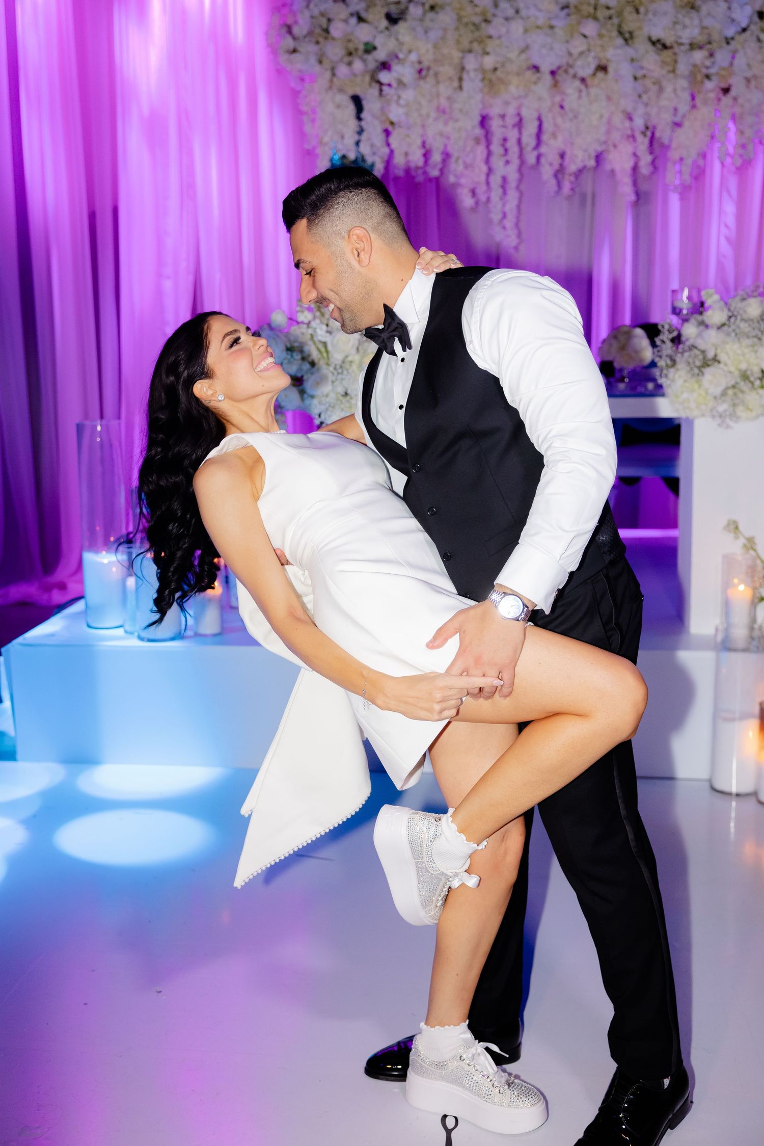 Couple in formal attire dancing intimately at purple-lit reception with floral backdrop.