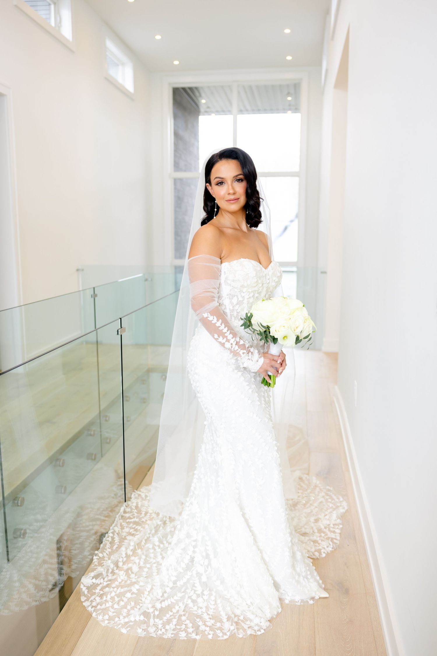 A bride in a white lace wedding dress with a long train holds a bouquet while standing in a modern hallway.