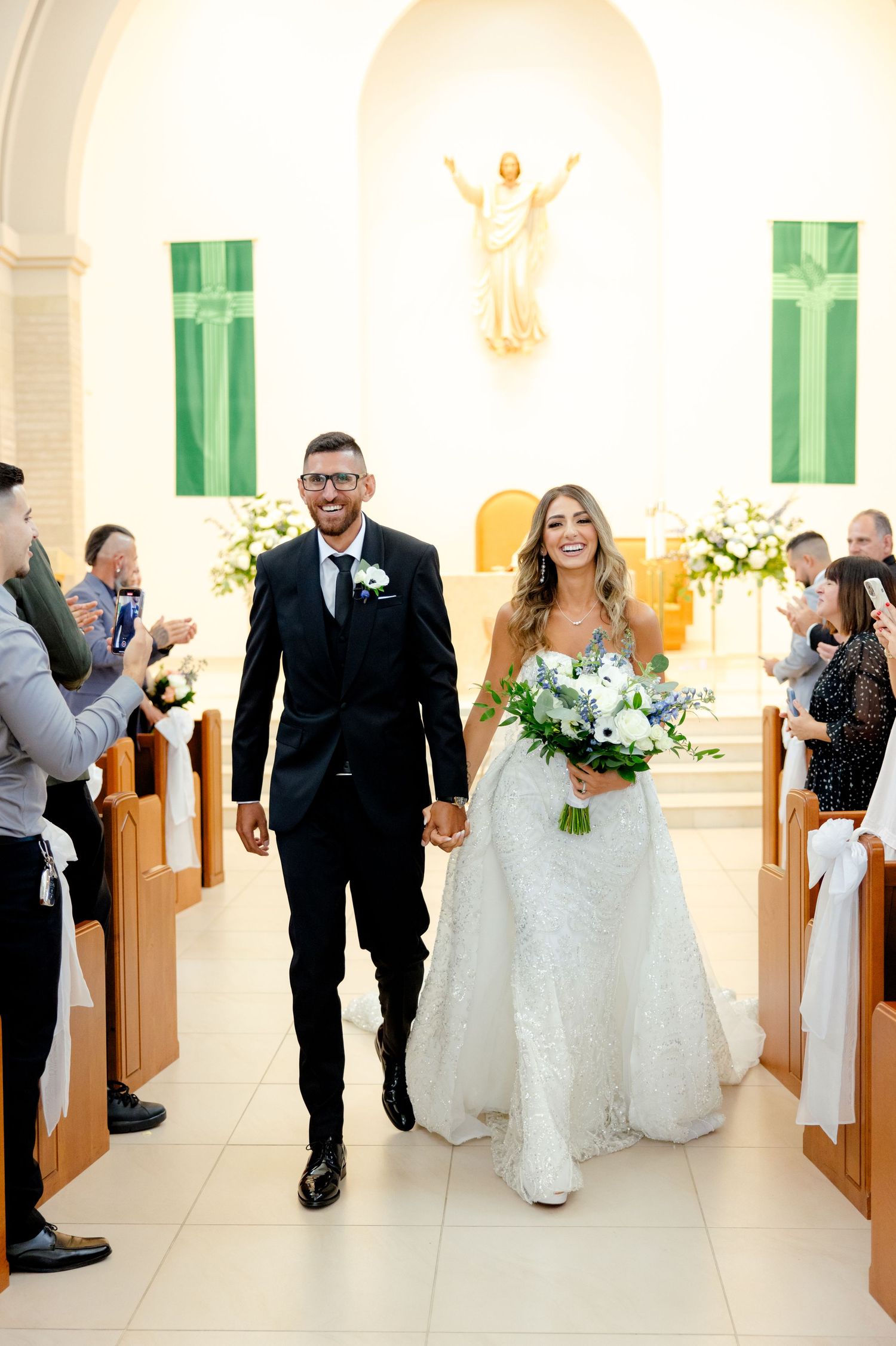 Happy newlyweds walking down church aisle while guests applaud from wooden pews.